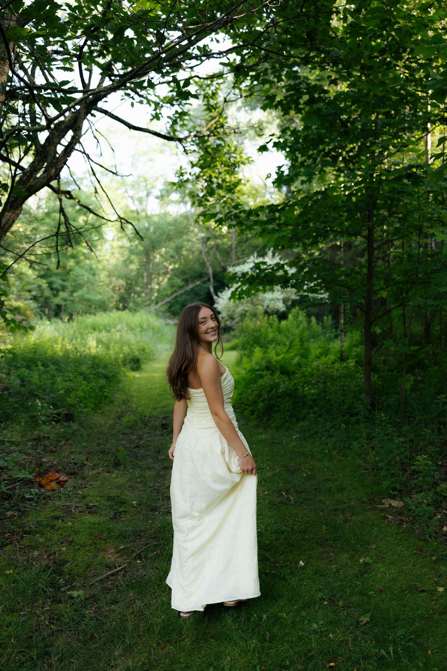 Young woman in white strapless gown smiling in lush green forest path.