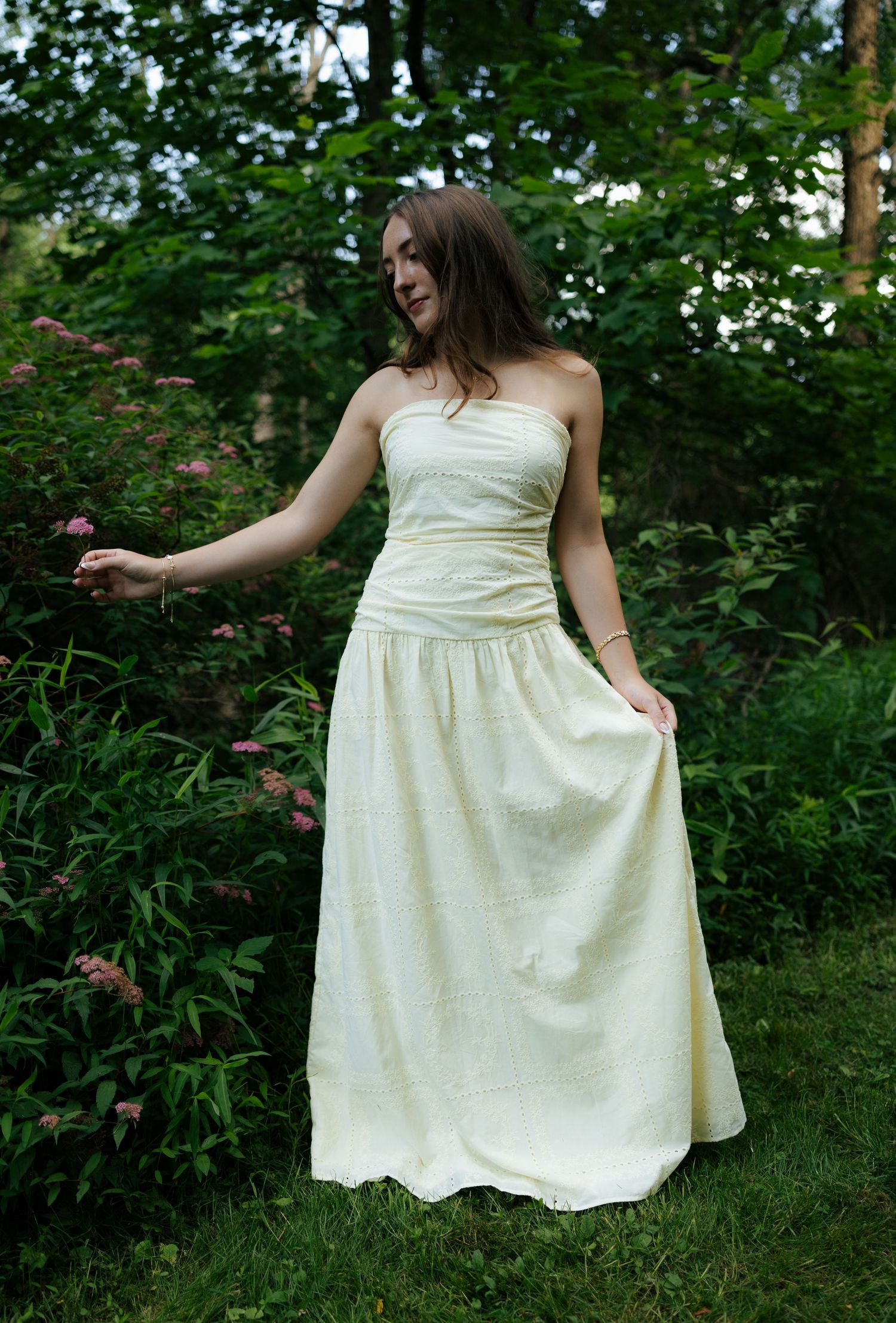 Young woman in cream strapless maxi dress smiling among pink flowering shrubs in green garden.