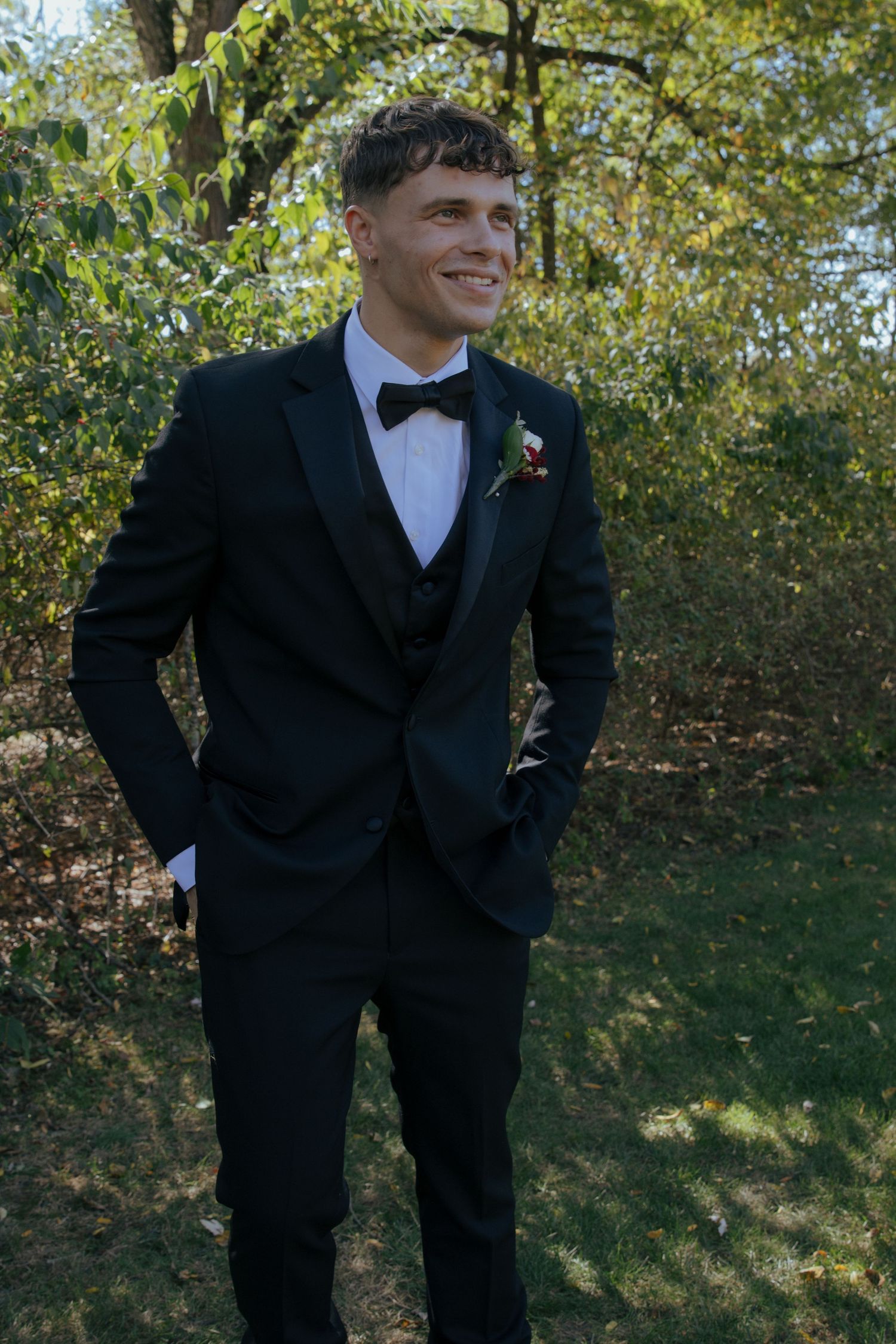 Young man in black tuxedo with bow tie and boutonniere smiling outdoors among green trees.