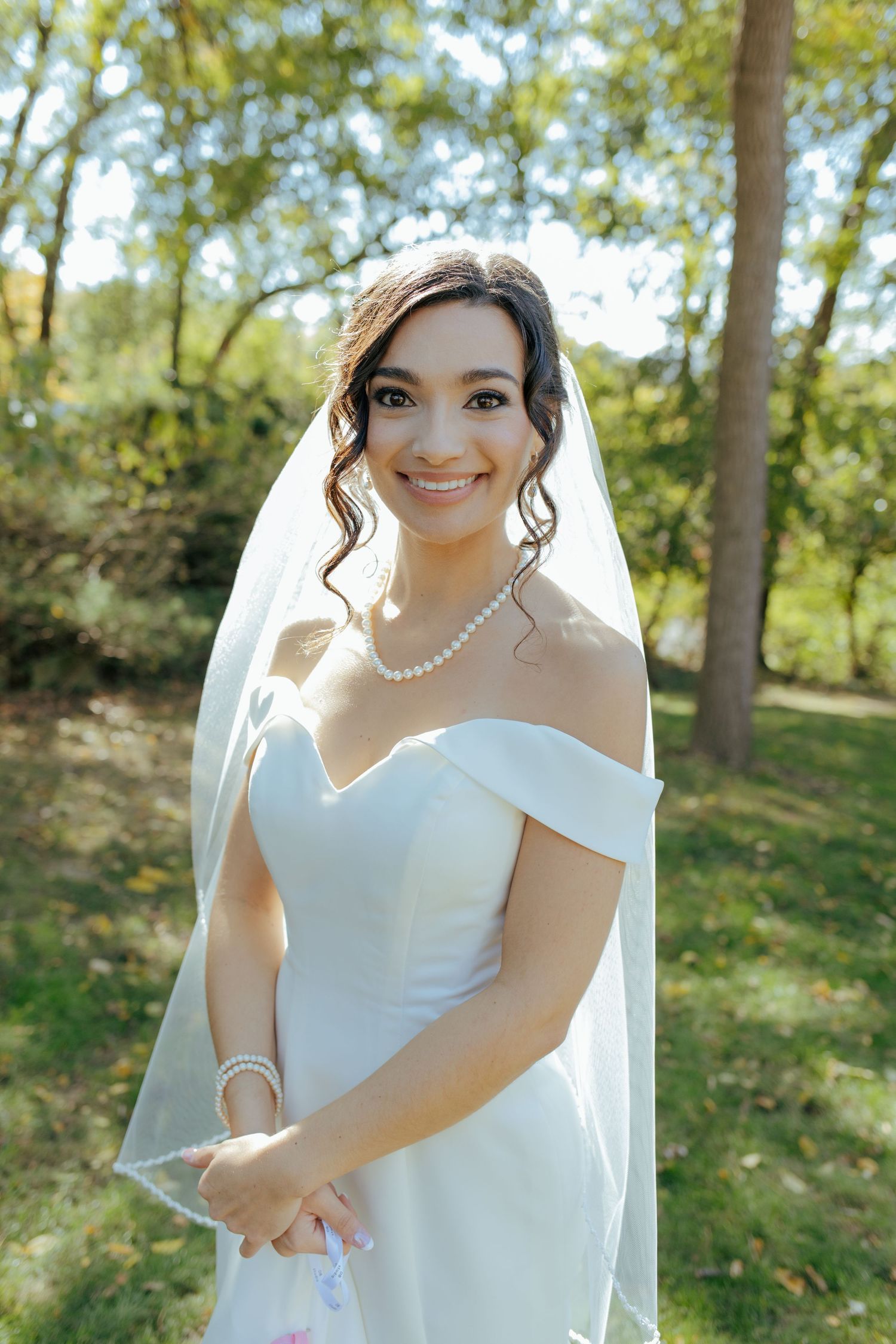 Radiant bride in off-shoulder white gown, pearl necklace, and veil smiles outdoors surrounded by green trees.