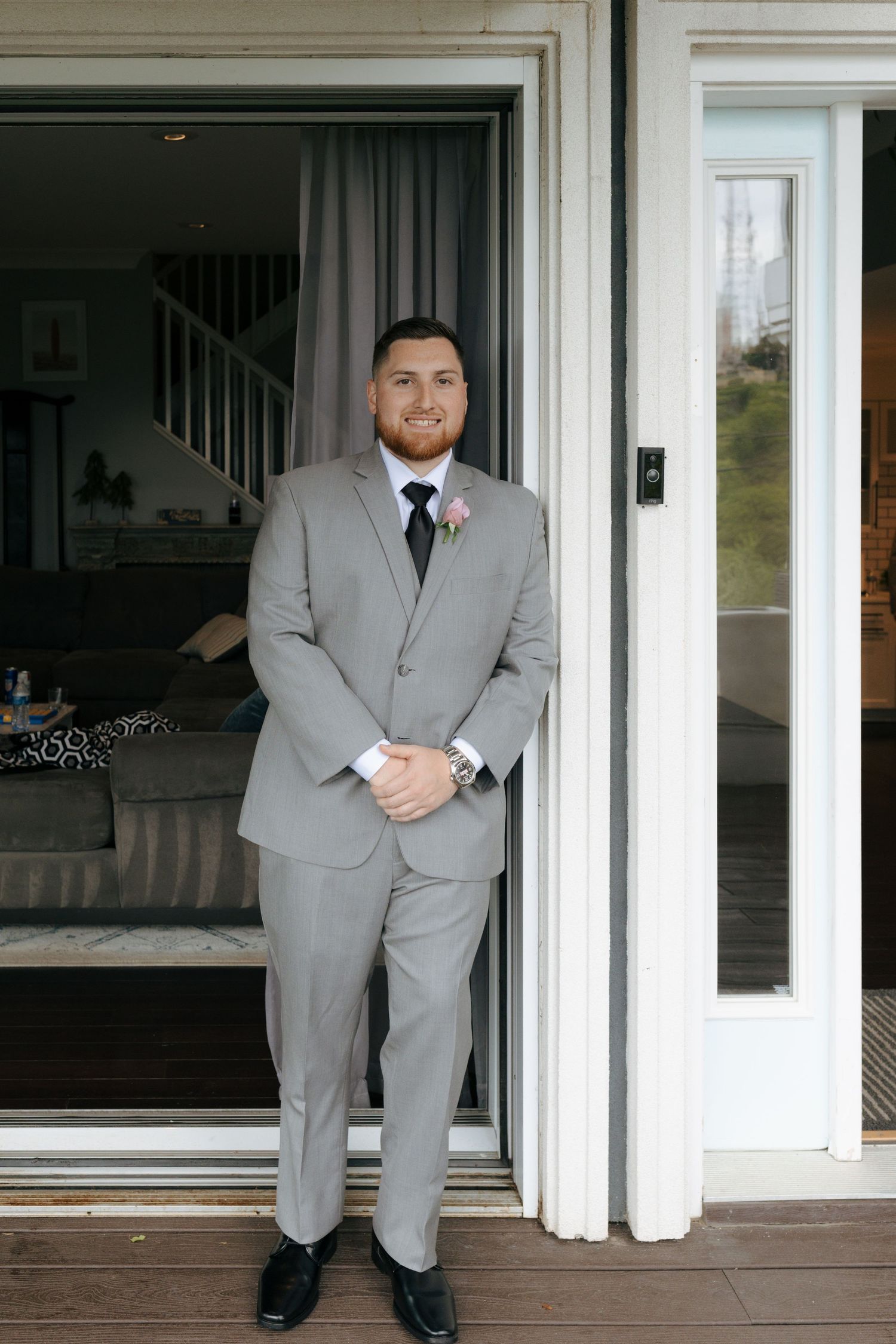 Groom in light gray suit with black tie and boutonniere stands in sliding door doorway on wedding day.