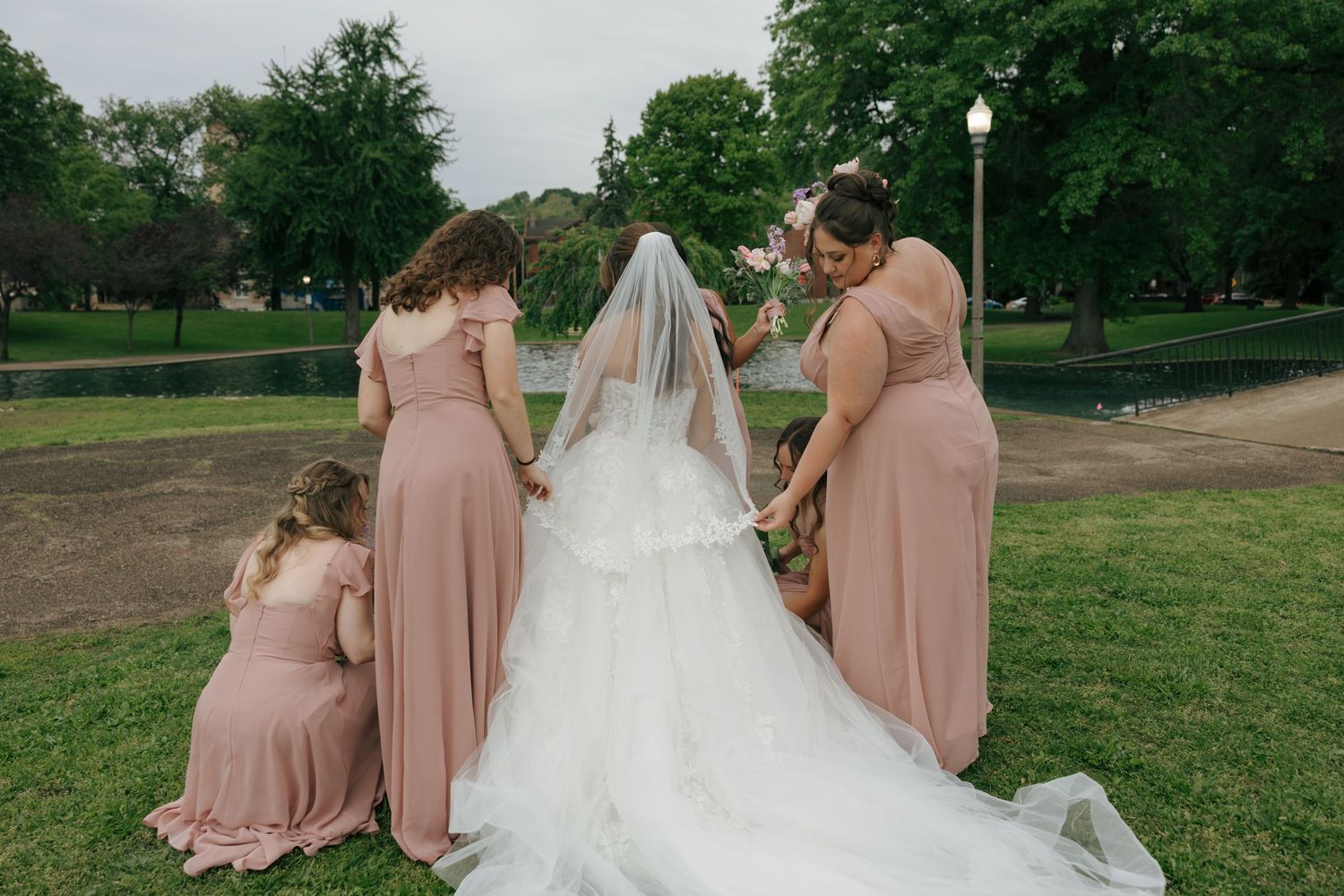 Bride in white gown and veil with bridesmaids in blush pink dresses in a lush green park setting.