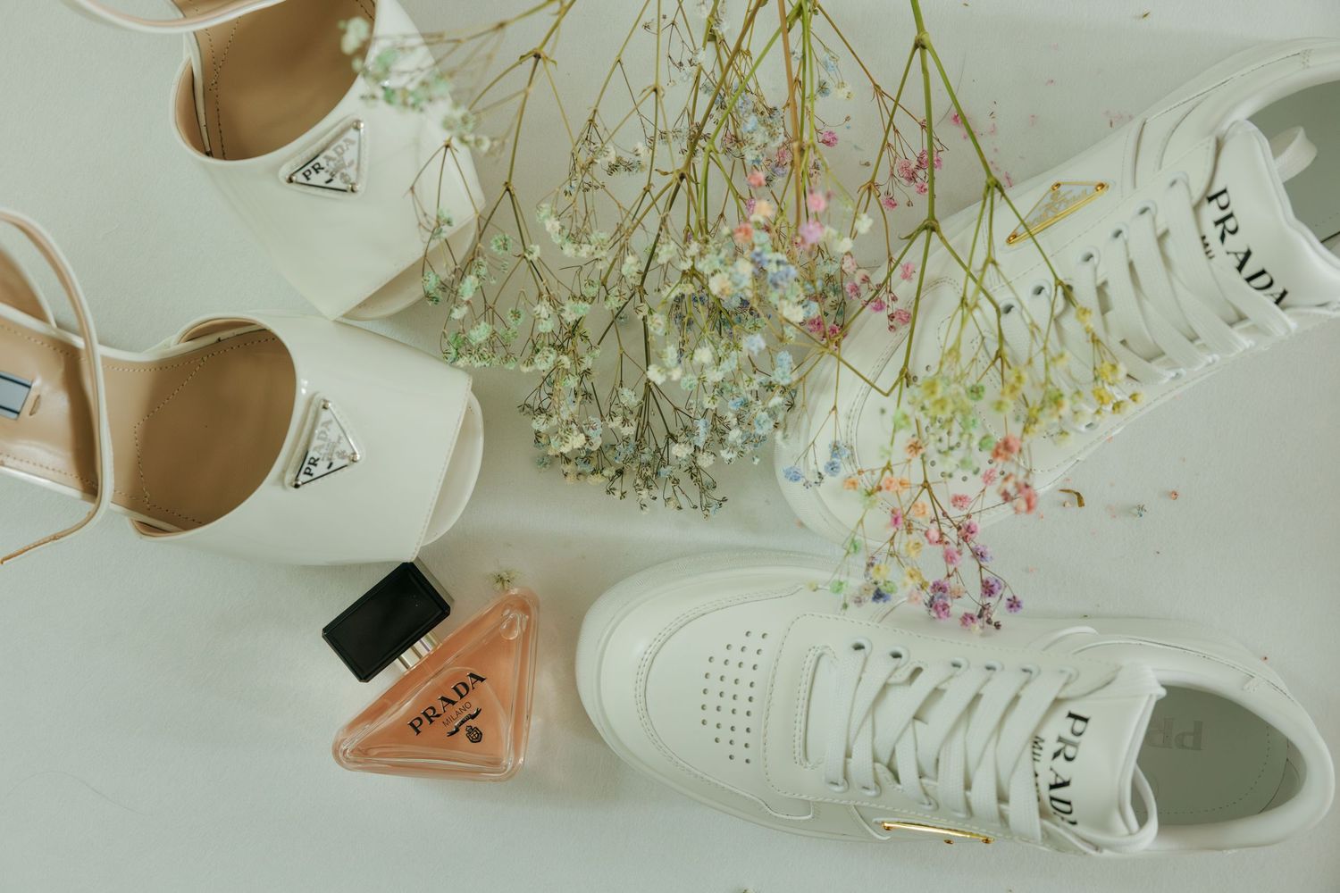 Prada white sneakers, heels, perfume bottle, and wildflowers arranged on a white surface.