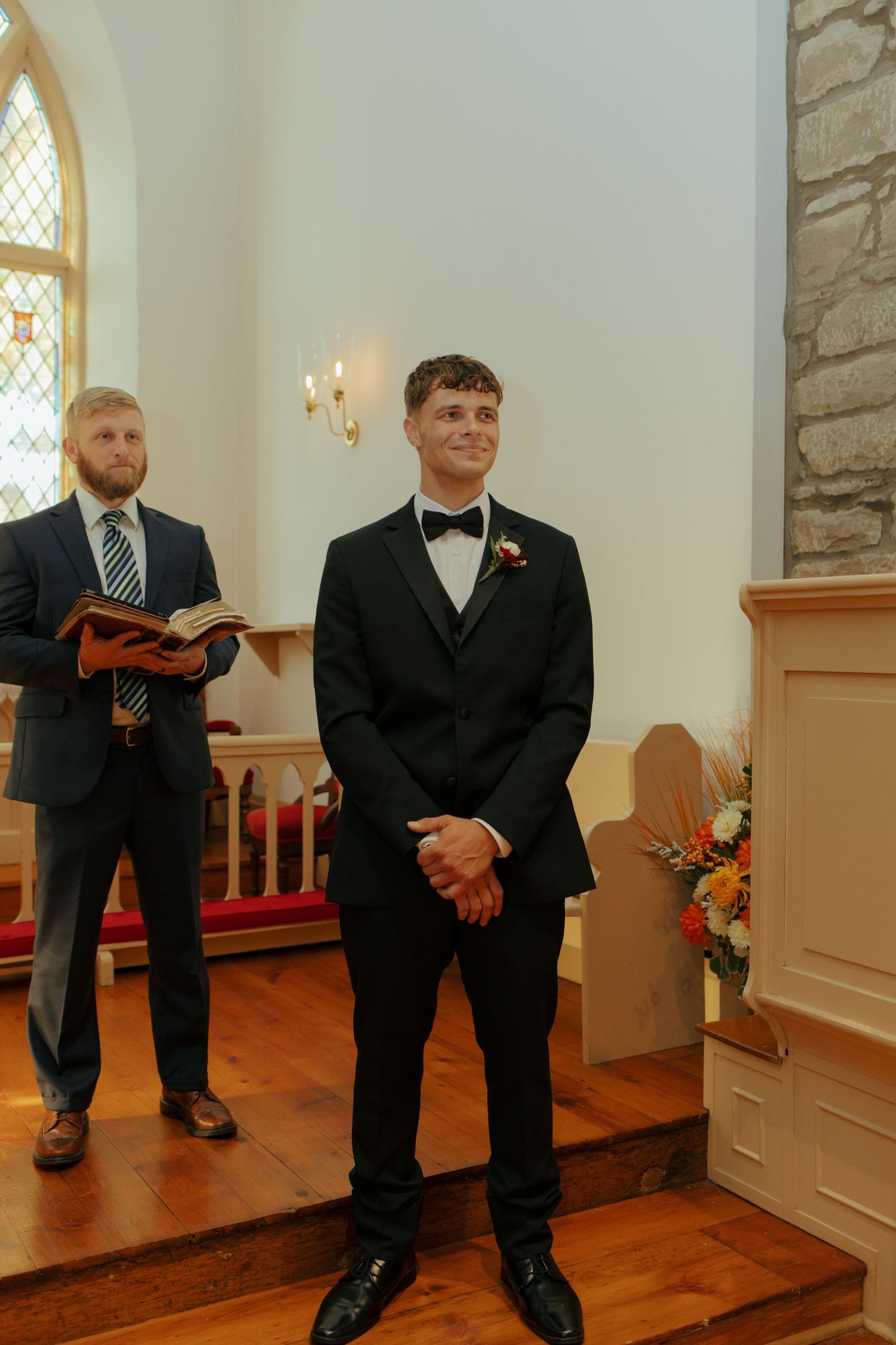 Groom in black tuxedo smiling at altar in stone church, officiant holding book behind him.