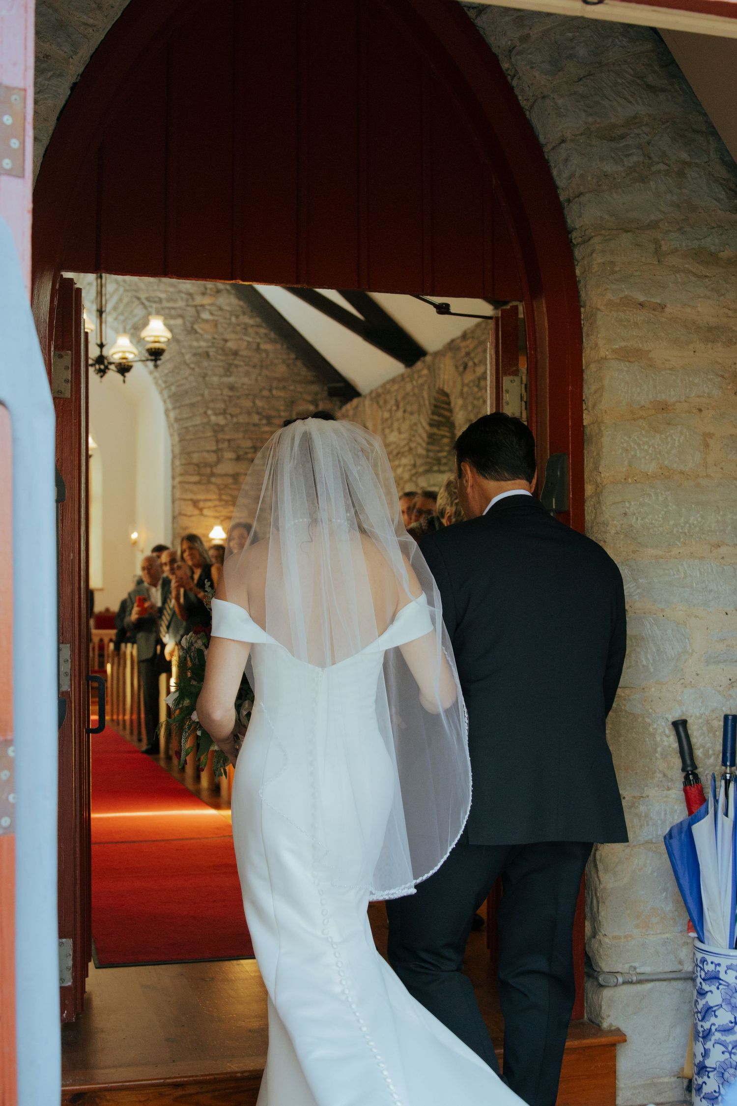 Bride in white gown and veil enters a stone church through an arched wooden doorway with escort.