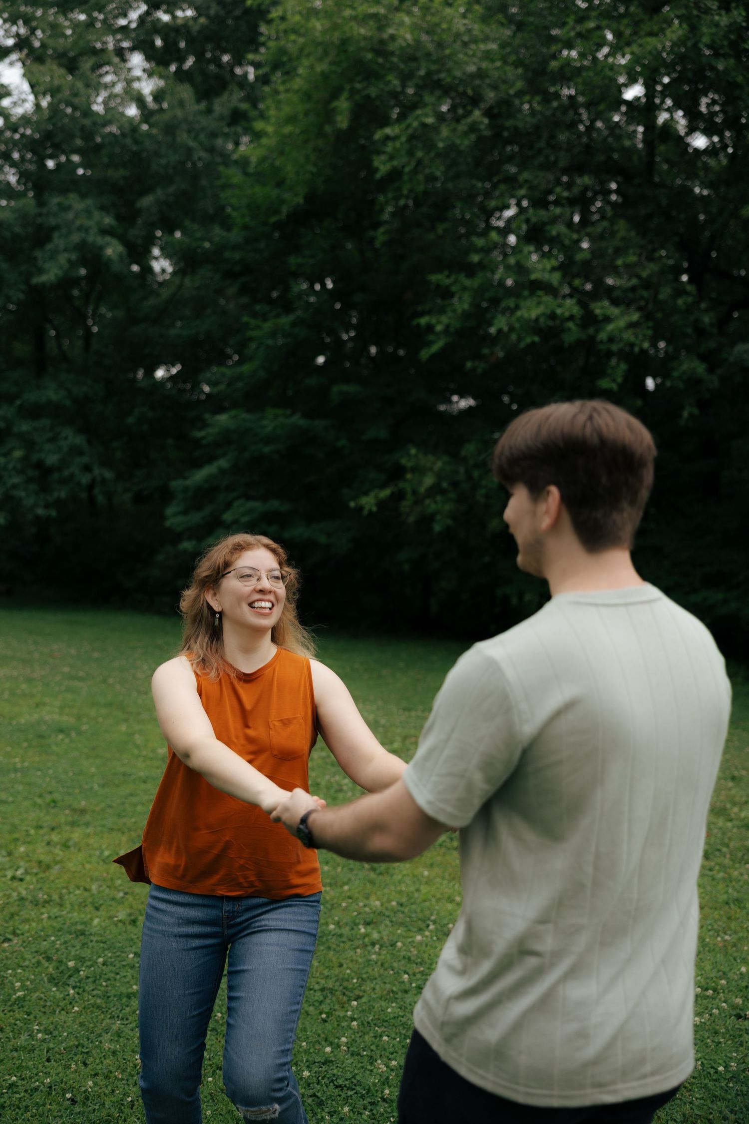 A laughing woman in an orange top holds hands with a man on a lush green lawn.