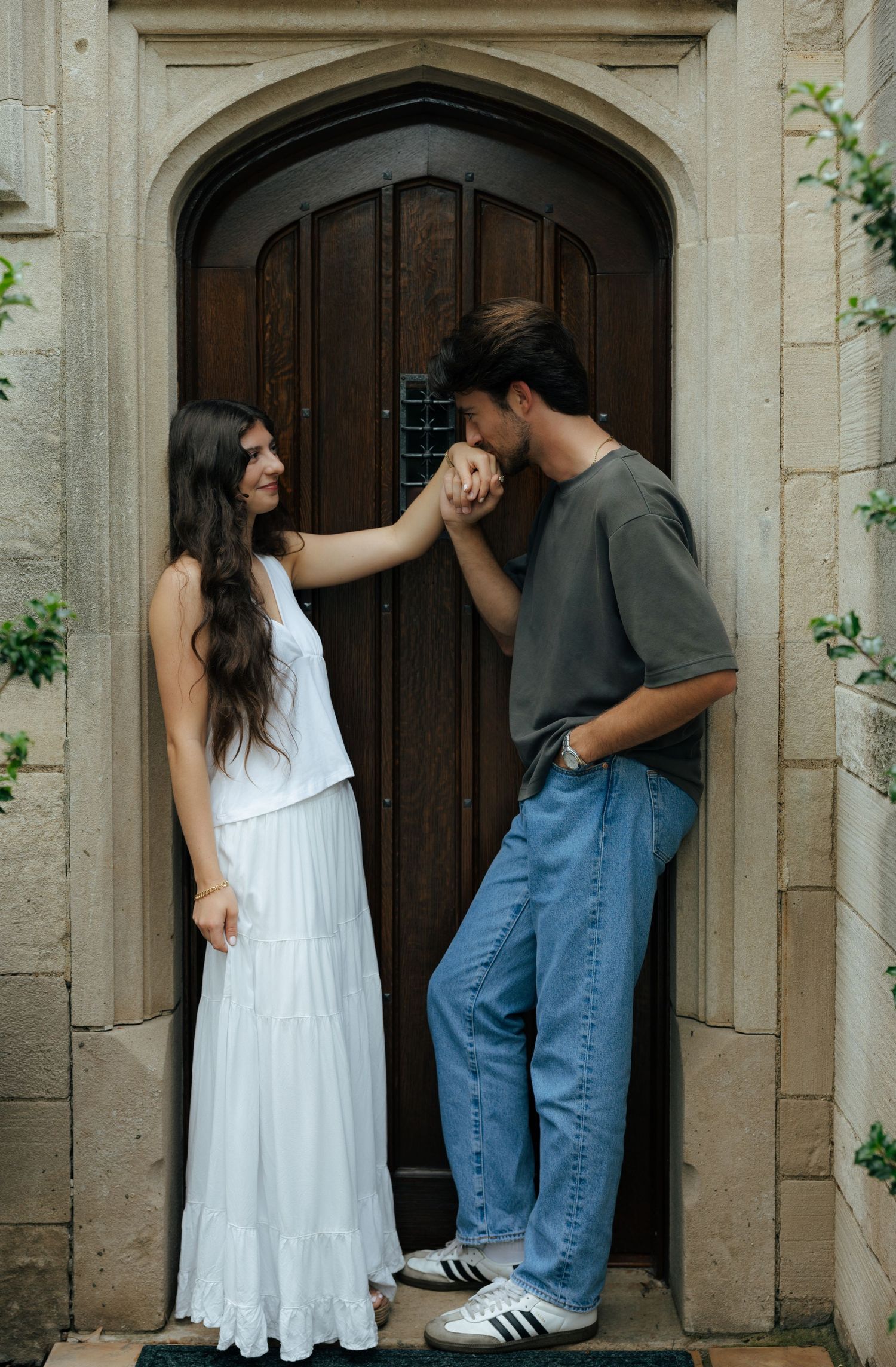 Couple sharing romantic hand-kiss moment before a gothic arched wooden door, woman in white tiered maxi dress.