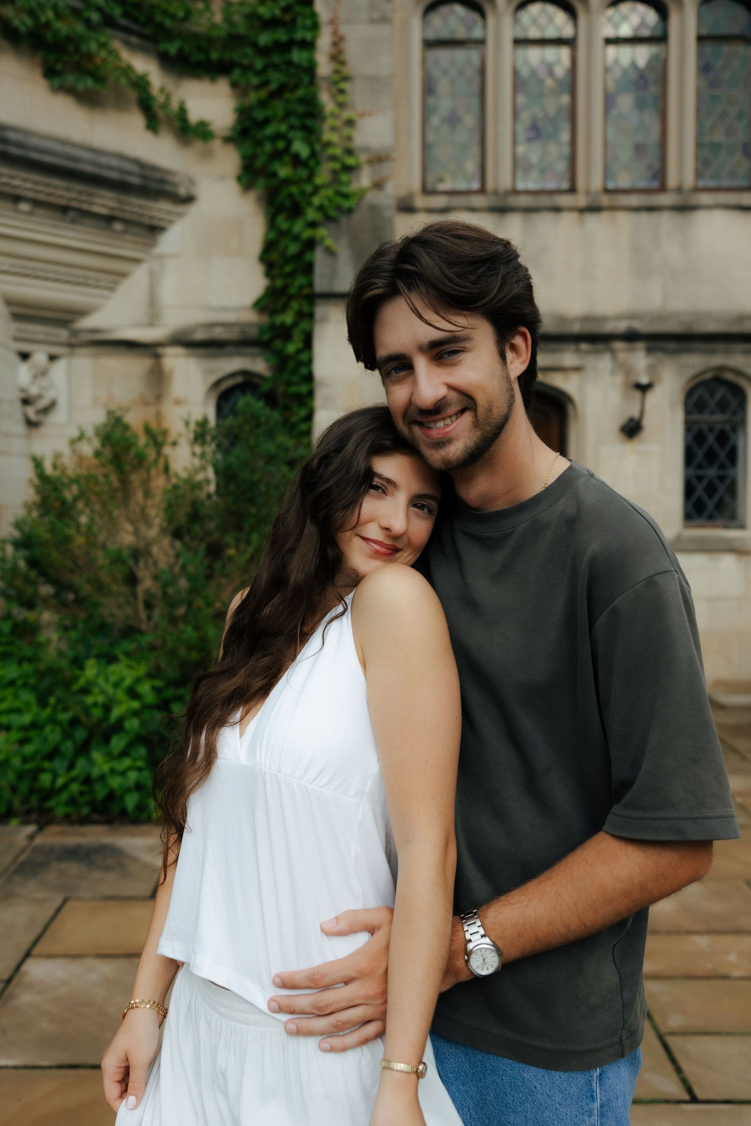 Smiling engaged couple embracing in front of ivy-covered gothic stone building during engagement session.