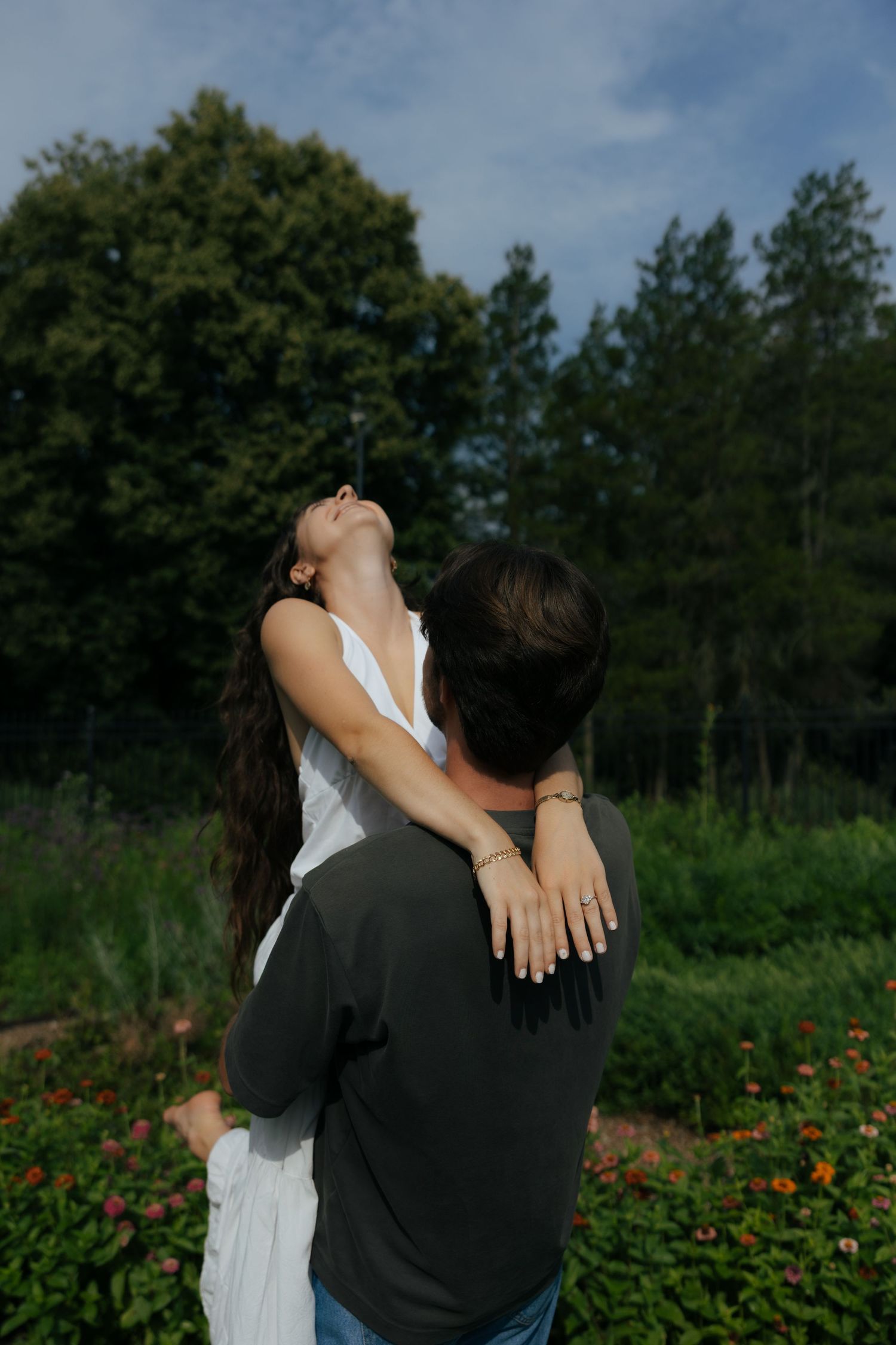 A man lifts a woman in a white dress as she leans back joyfully in a lush garden.