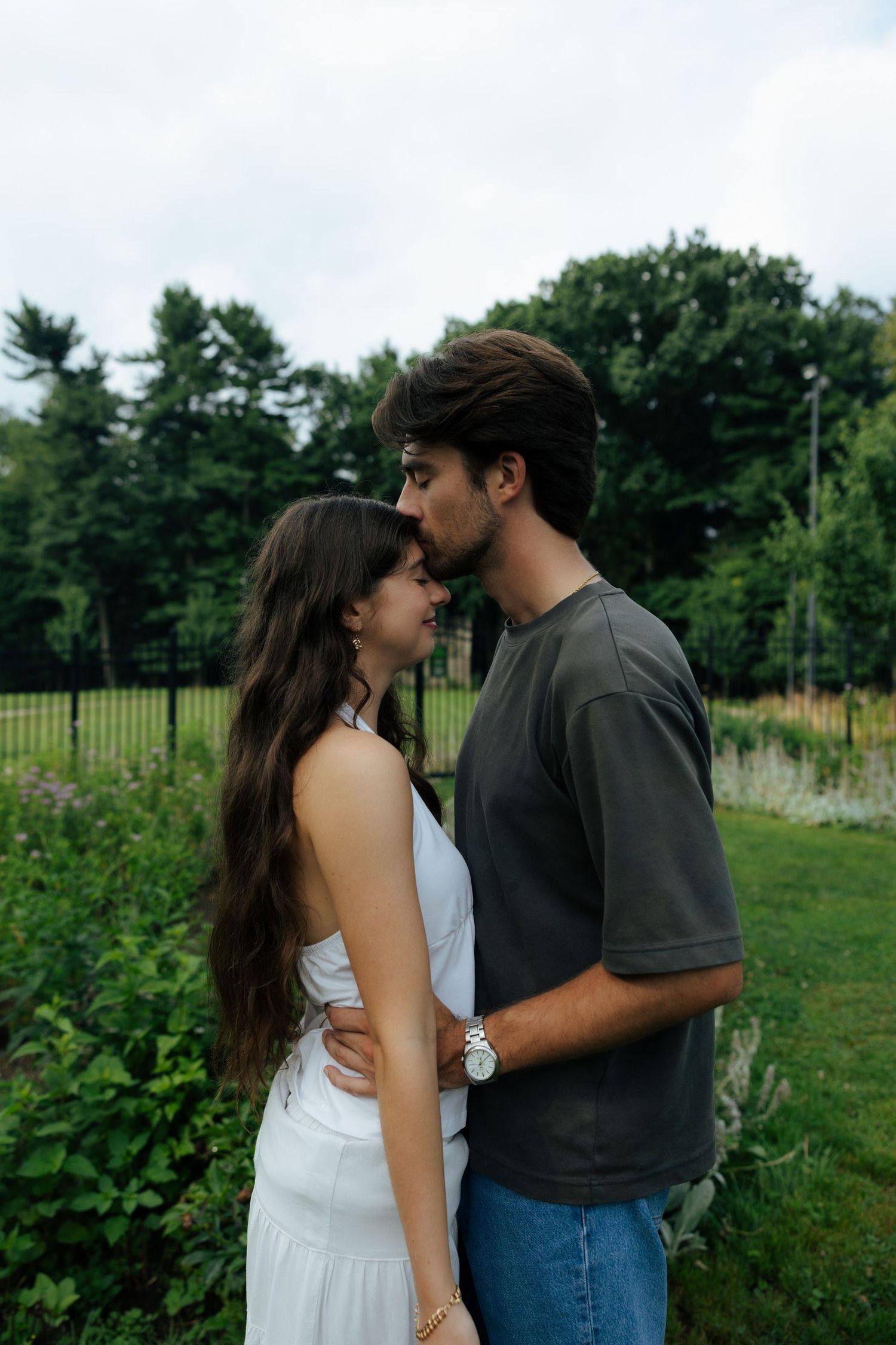 A young couple shares an intimate moment outdoors, nearly kissing while embracing in a lush green park.