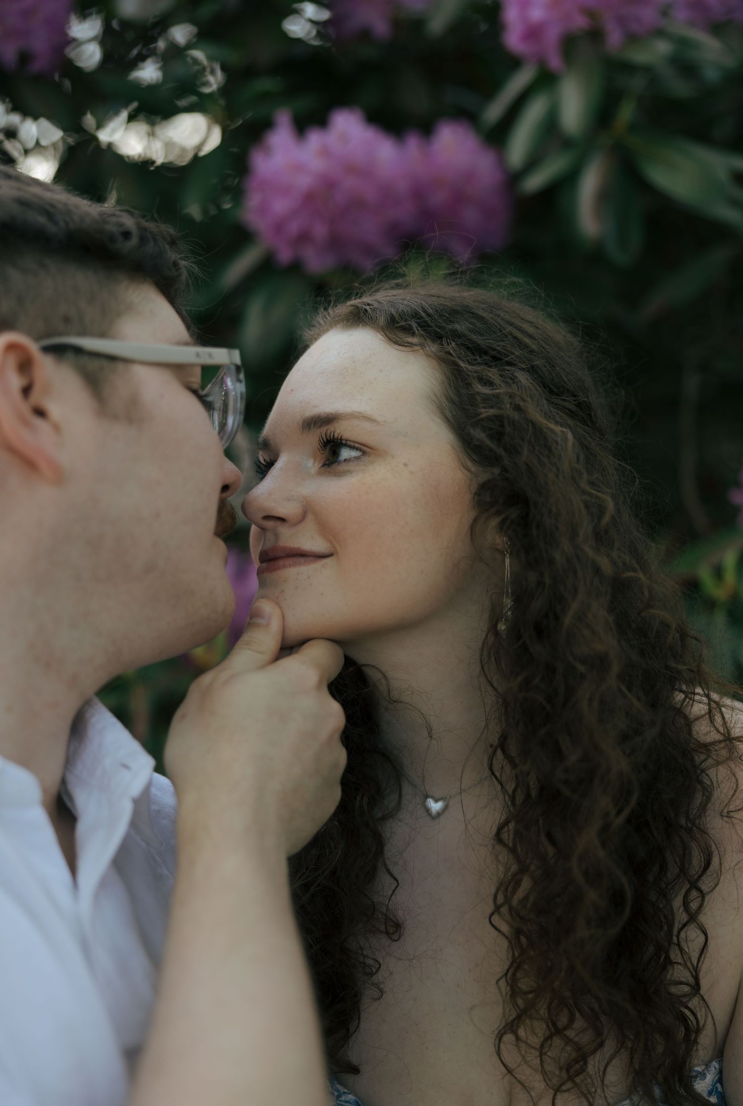 Man gently holding woman's chin near blooming pink rhododendrons, woman smiling softly during outdoor engagement shoot.