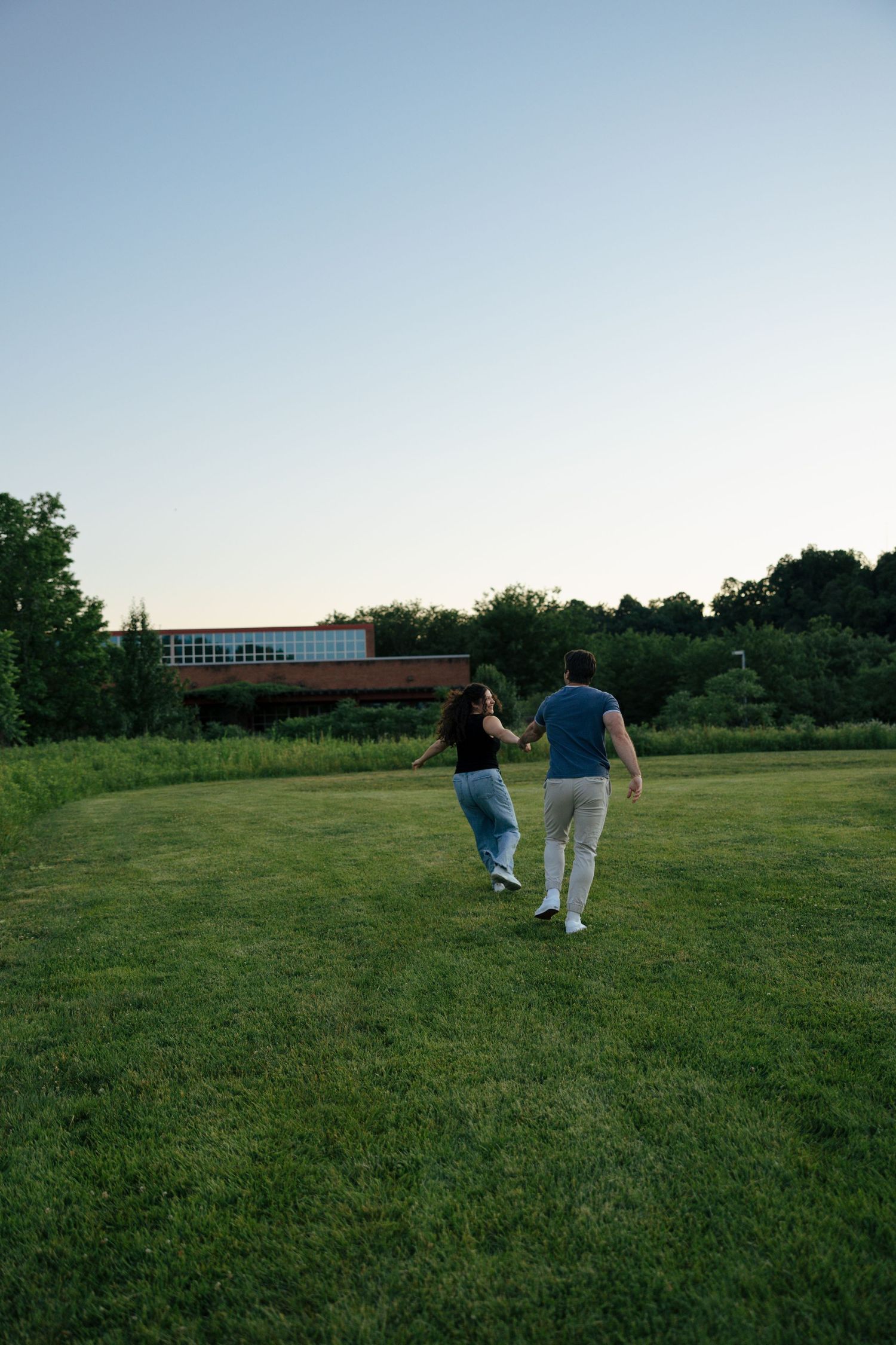 A couple holds hands while walking across a lush green lawn at sunset with trees in the background.