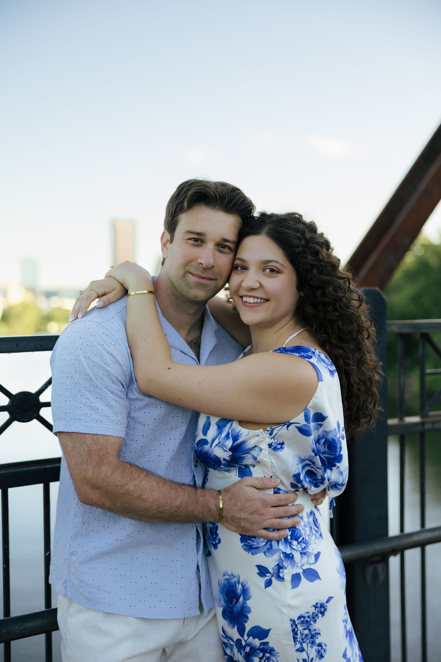 A smiling couple hugs affectionately on a bridge with a city skyline and blue sky behind them.