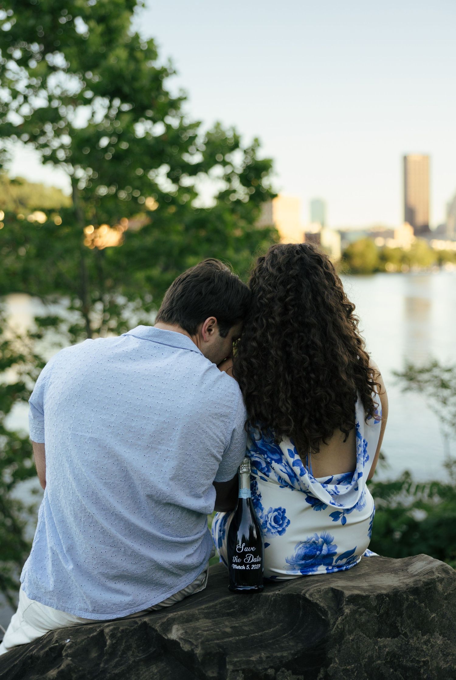 Couple sitting together on rocks overlooking a city skyline river view with save-the-date bottles.