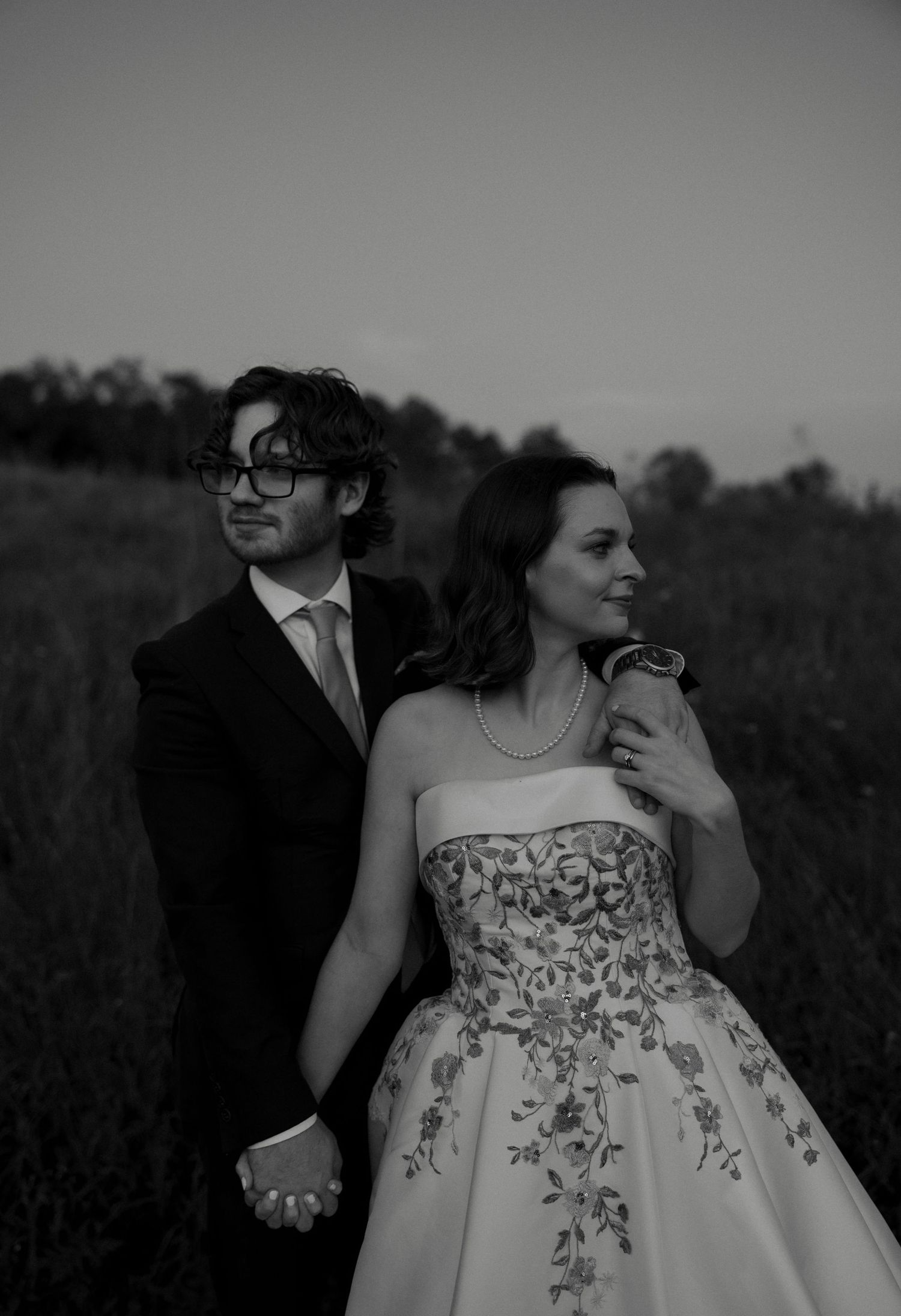 Black and white photo of bride in floral gown and groom in suit holding hands in a field at dusk.