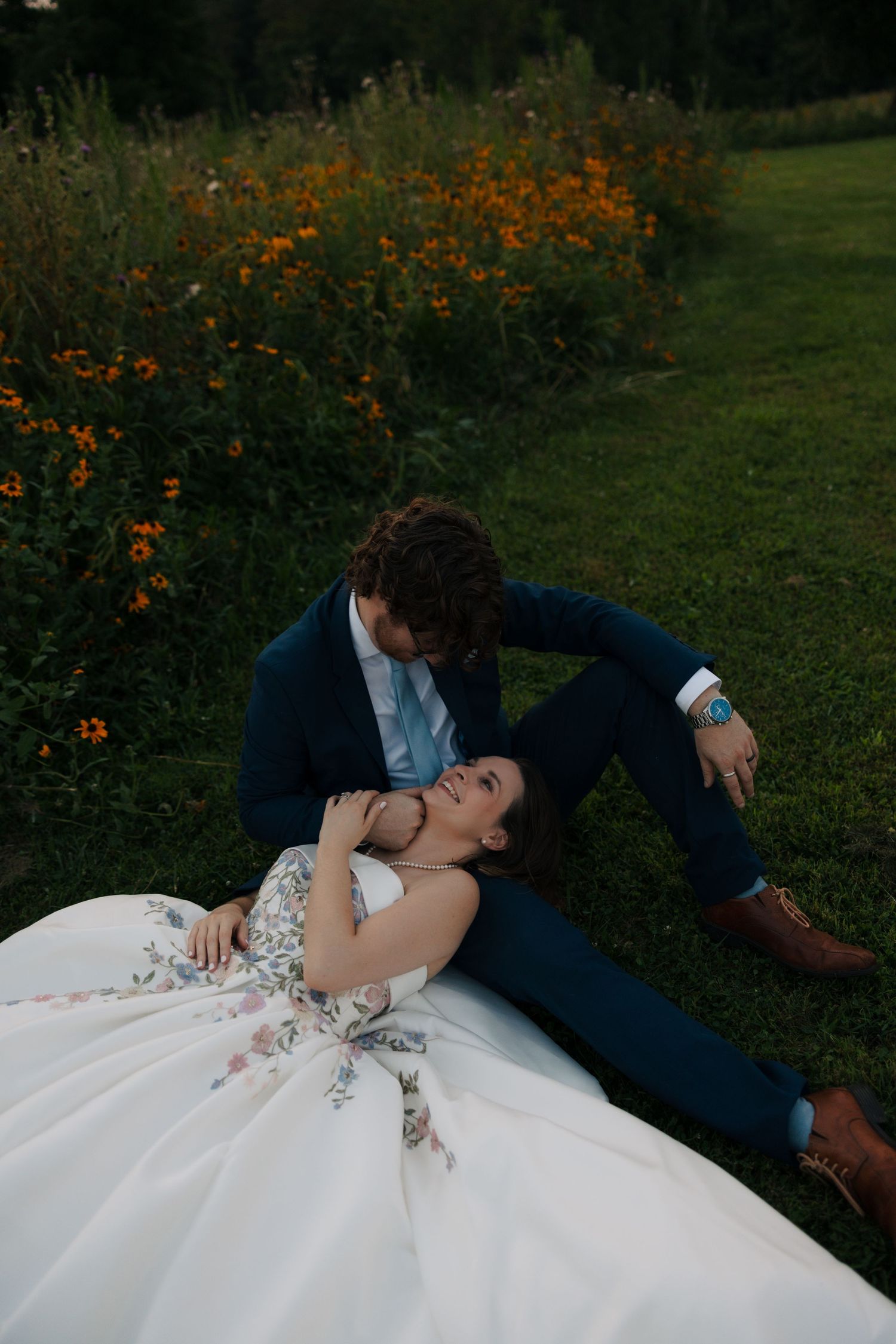 Bride in floral gown reclines on groom's lap while sitting on grass beside vibrant orange wildflowers.