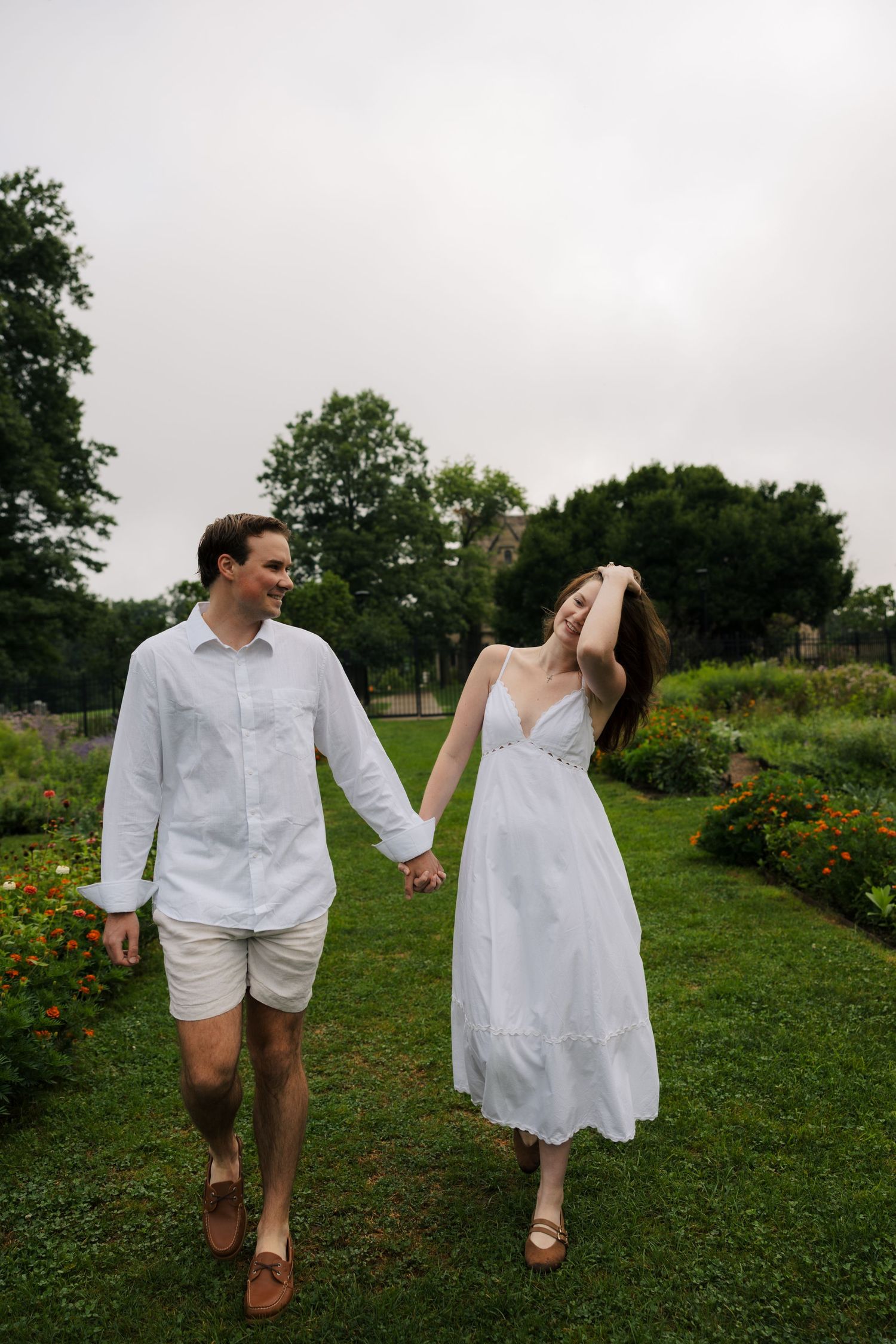 A smiling couple in white outfits walk hand-in-hand through a lush green garden on an overcast day.