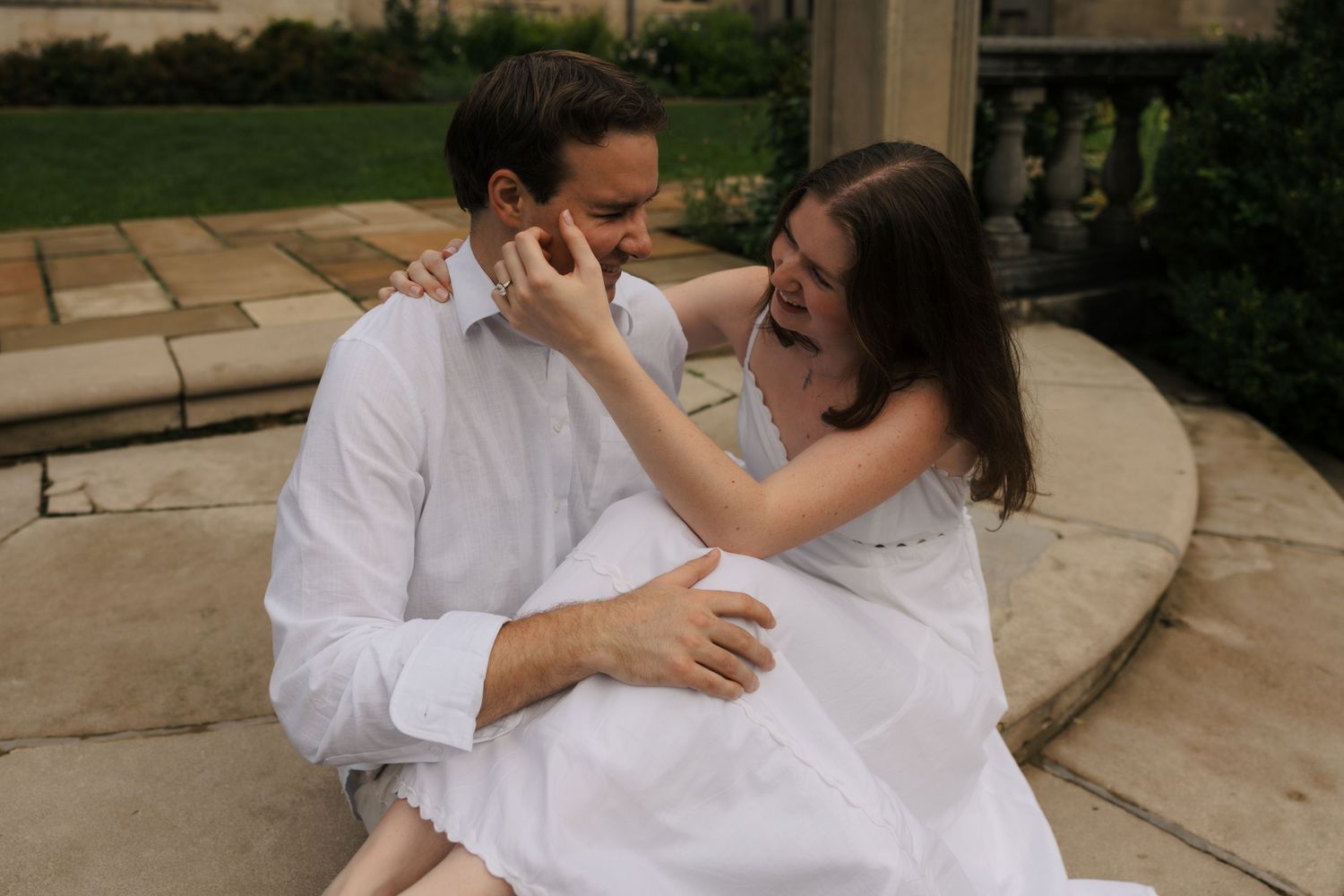 Laughing couple sitting on stone steps, woman tenderly touching man's face during engagement shoot.