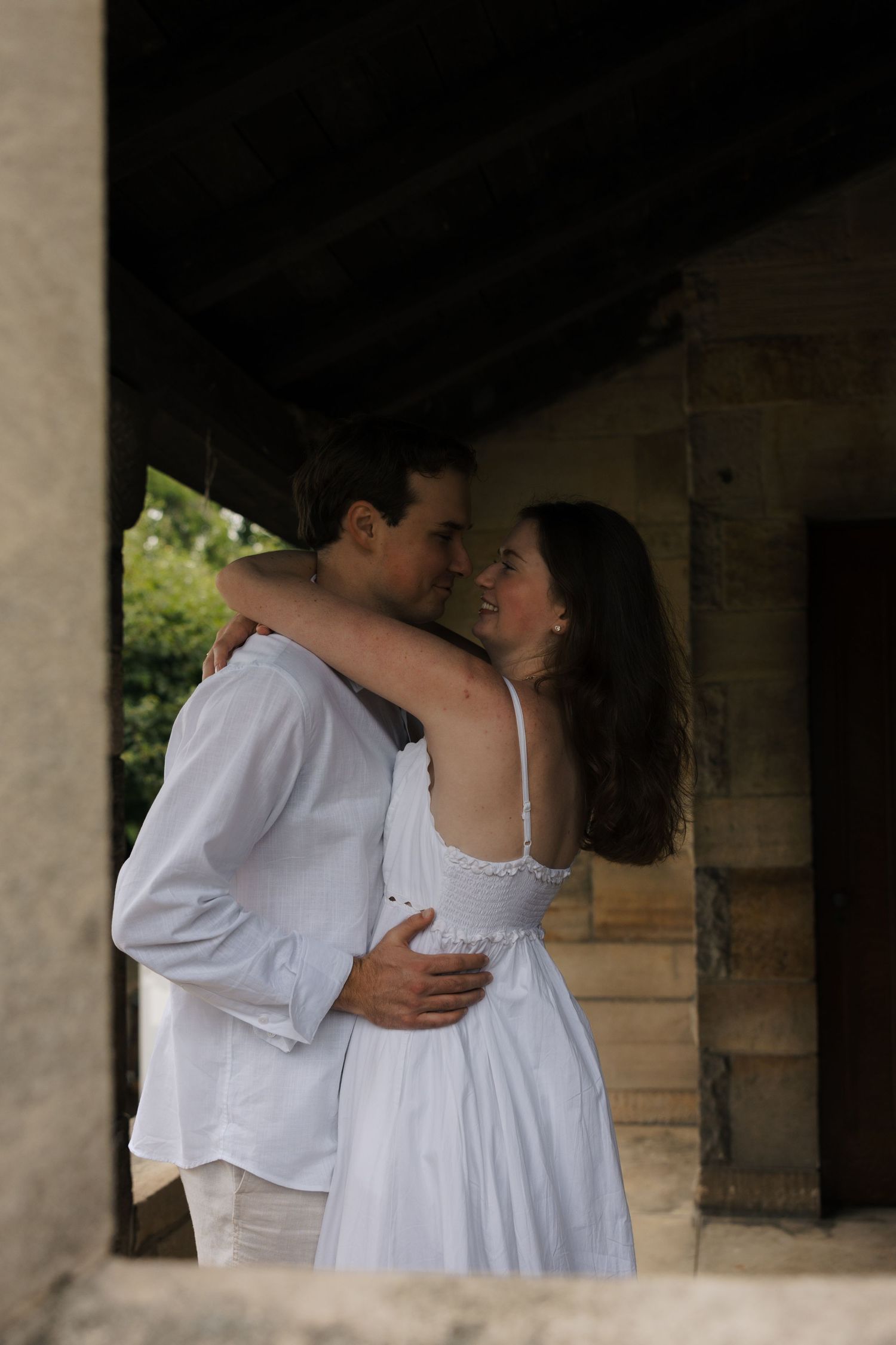 Couple in white attire embraces and smiles in shadowed stone archway during engagement shoot.