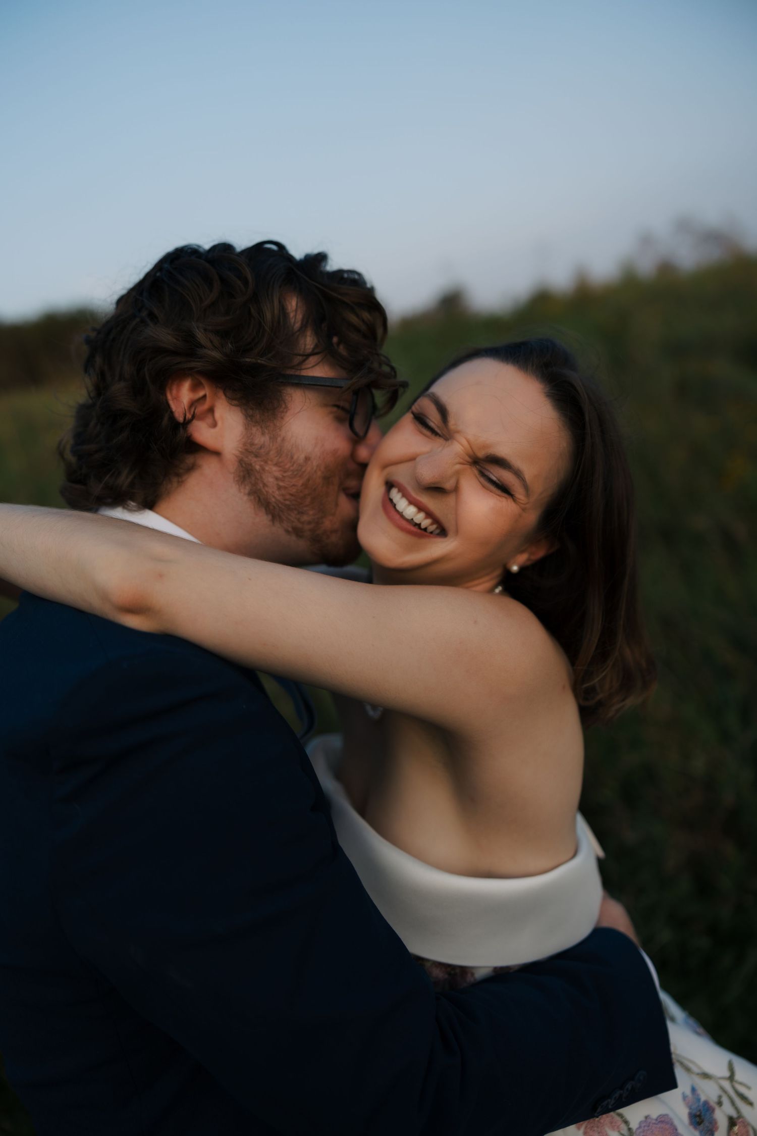 Bride laughs joyfully as groom kisses her cheek during romantic outdoor golden hour portraits.