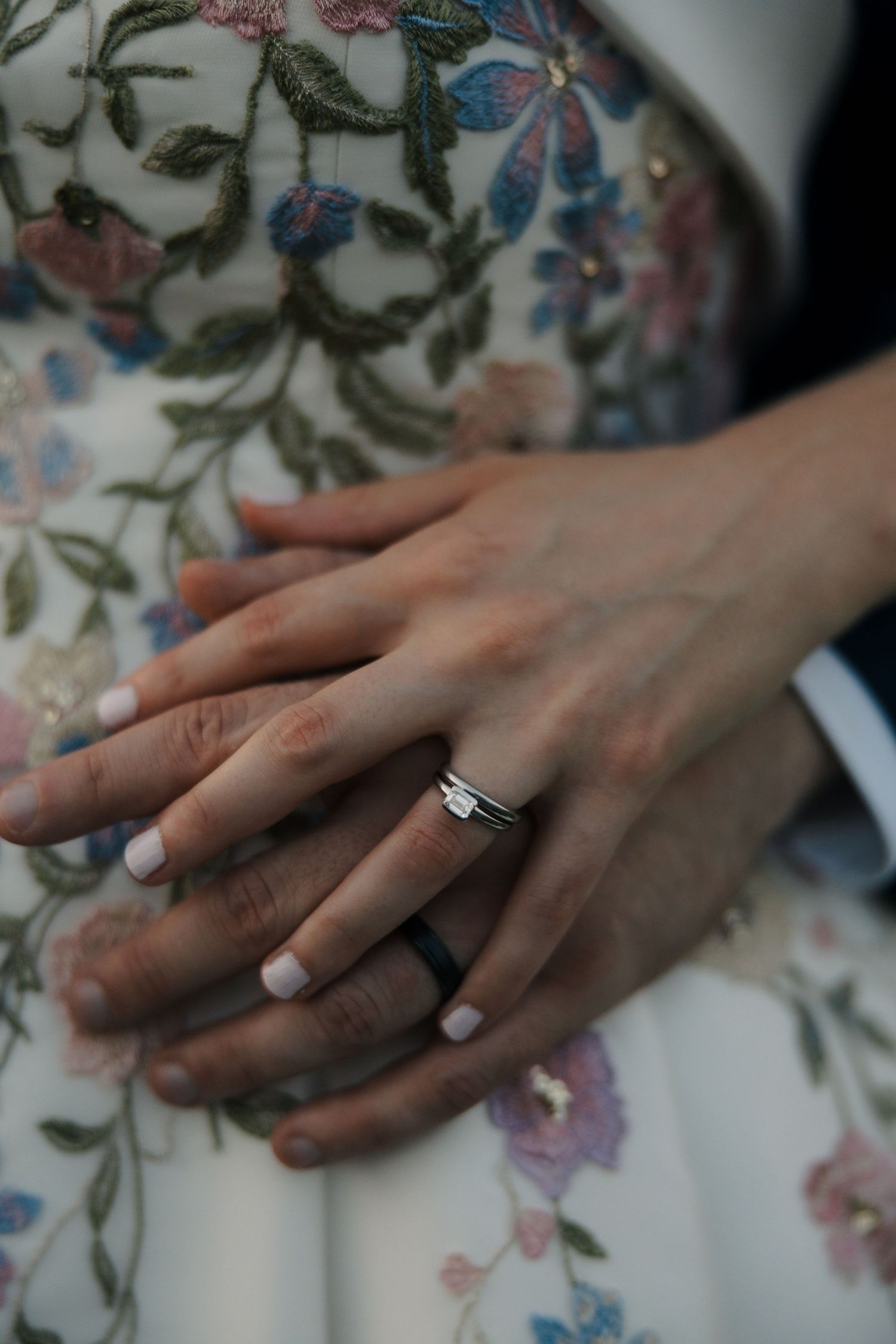 Close-up of bride and groom's hands showing wedding rings against intricate floral embroidered wedding dress.