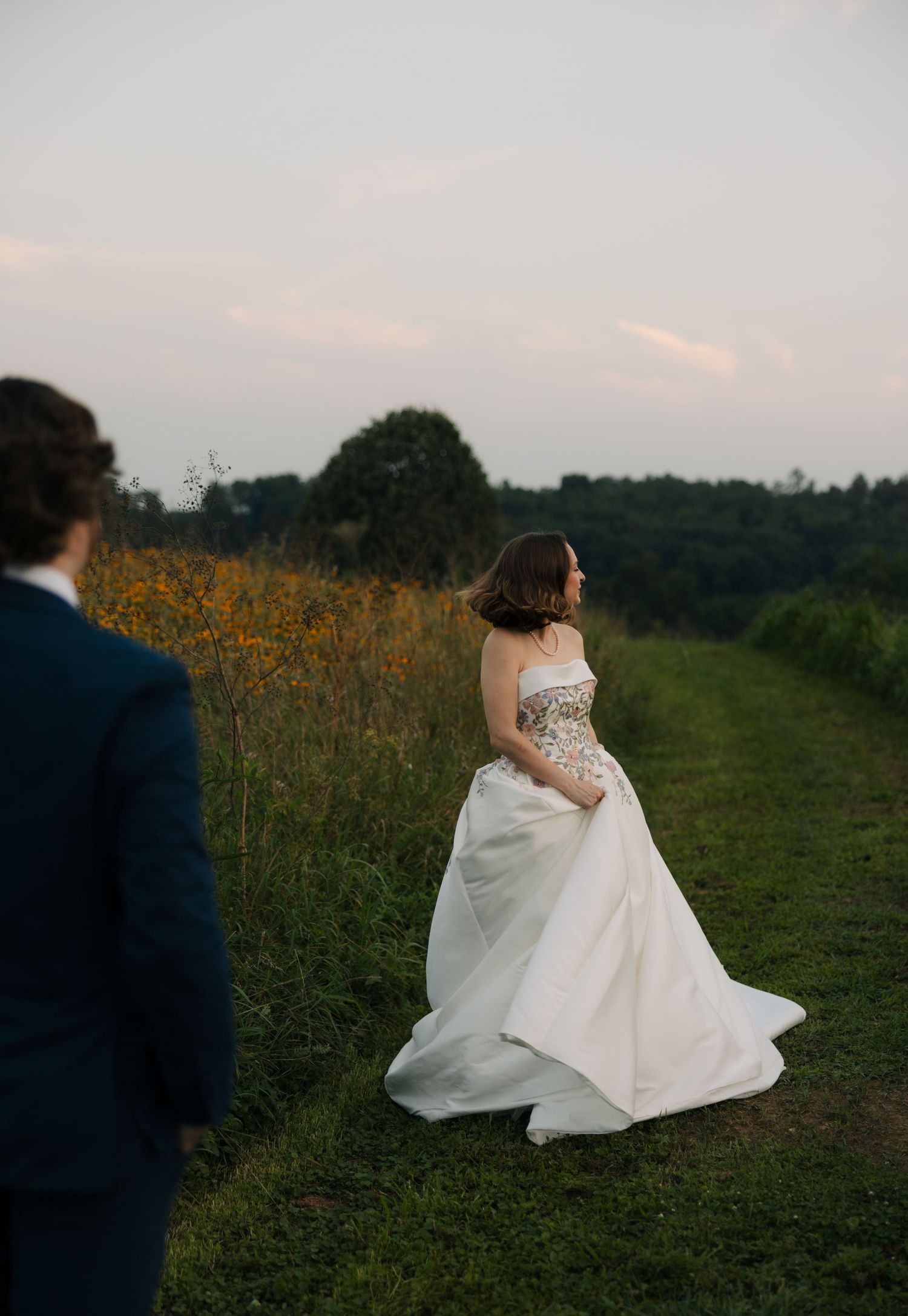 Bride in embellished strapless wedding gown stands on a grassy path surrounded by golden wildflowers at sunset.