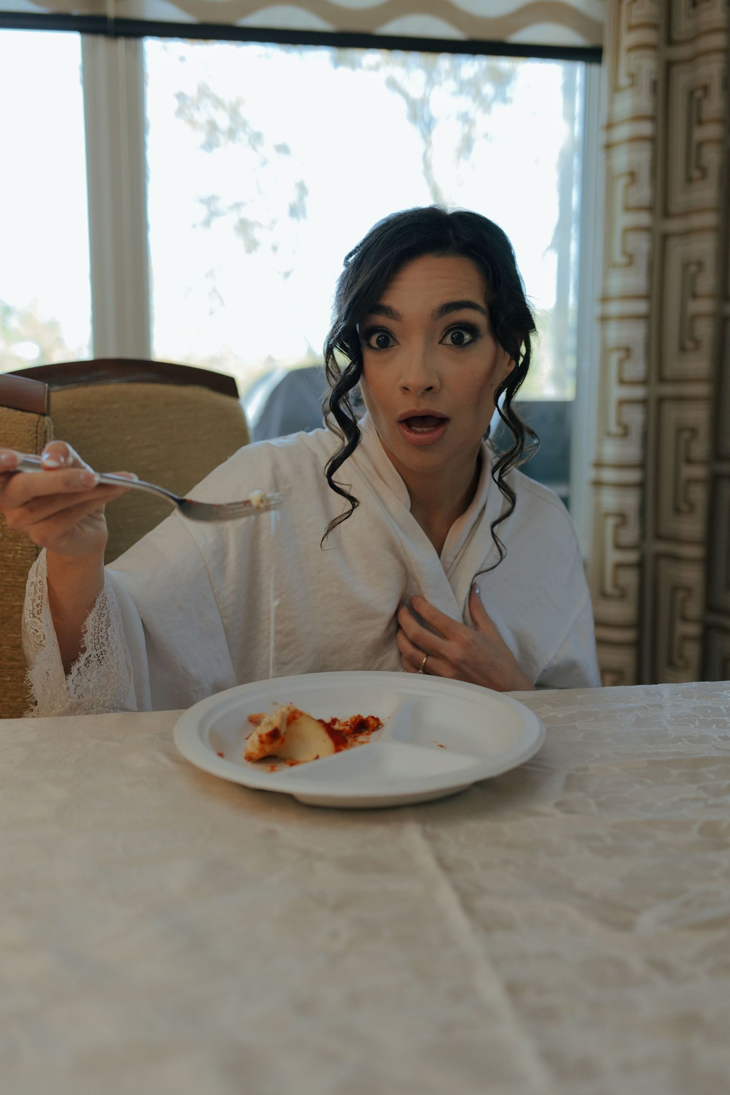 Surprised bride in white robe eating breakfast at a table before her wedding ceremony.