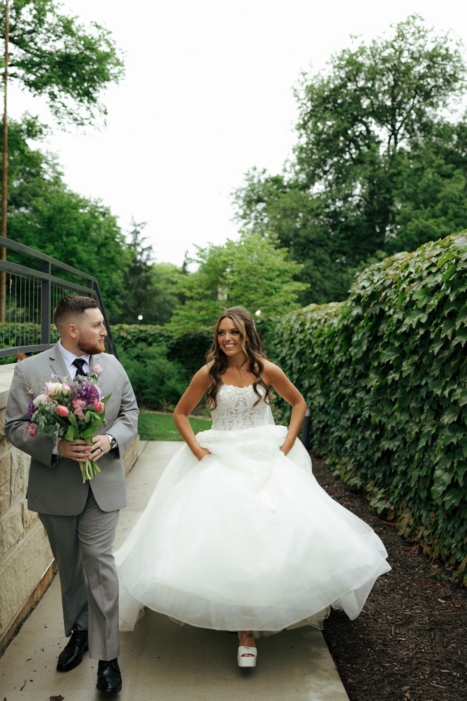 Bride in white ball gown and groom in gray suit walk together outdoors, surrounded by lush greenery on their wedding day.