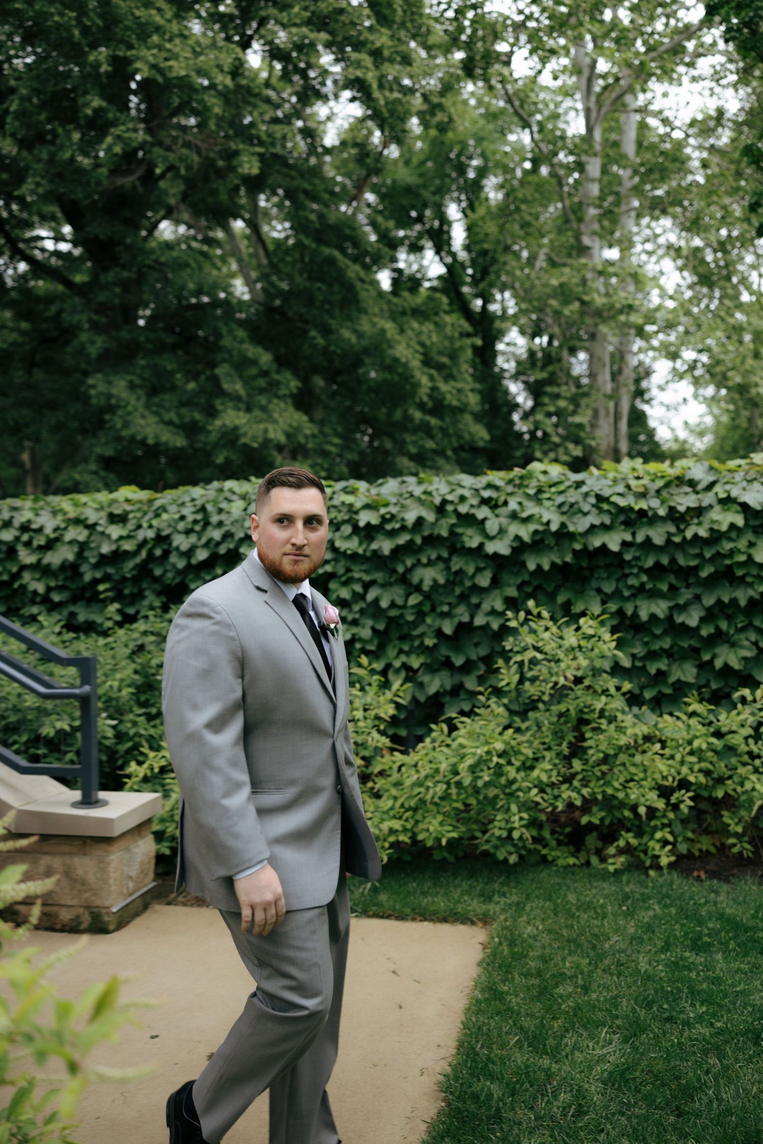 Groom in gray suit with boutonniere walks along garden path with lush green hedges at outdoor wedding.