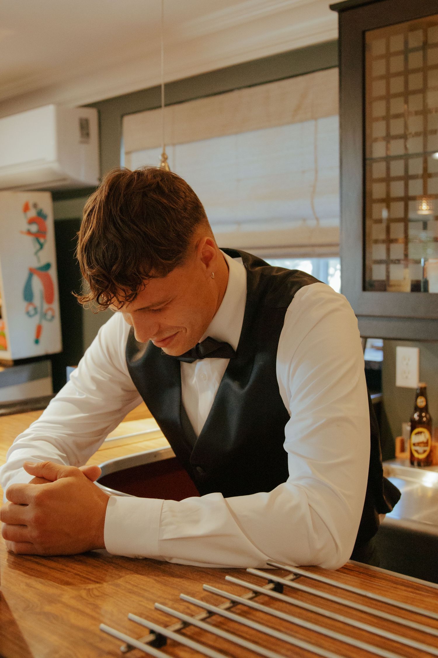 Bartender in black vest and bow tie leans on wooden bar counter at wedding venue.