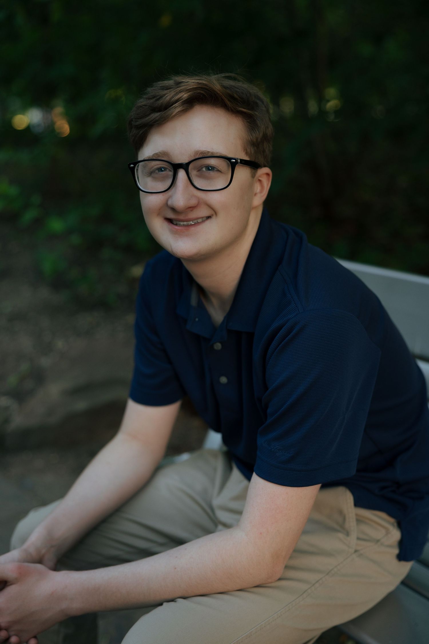 Young man with glasses smiling in navy polo shirt, seated outdoors with green trees in background.