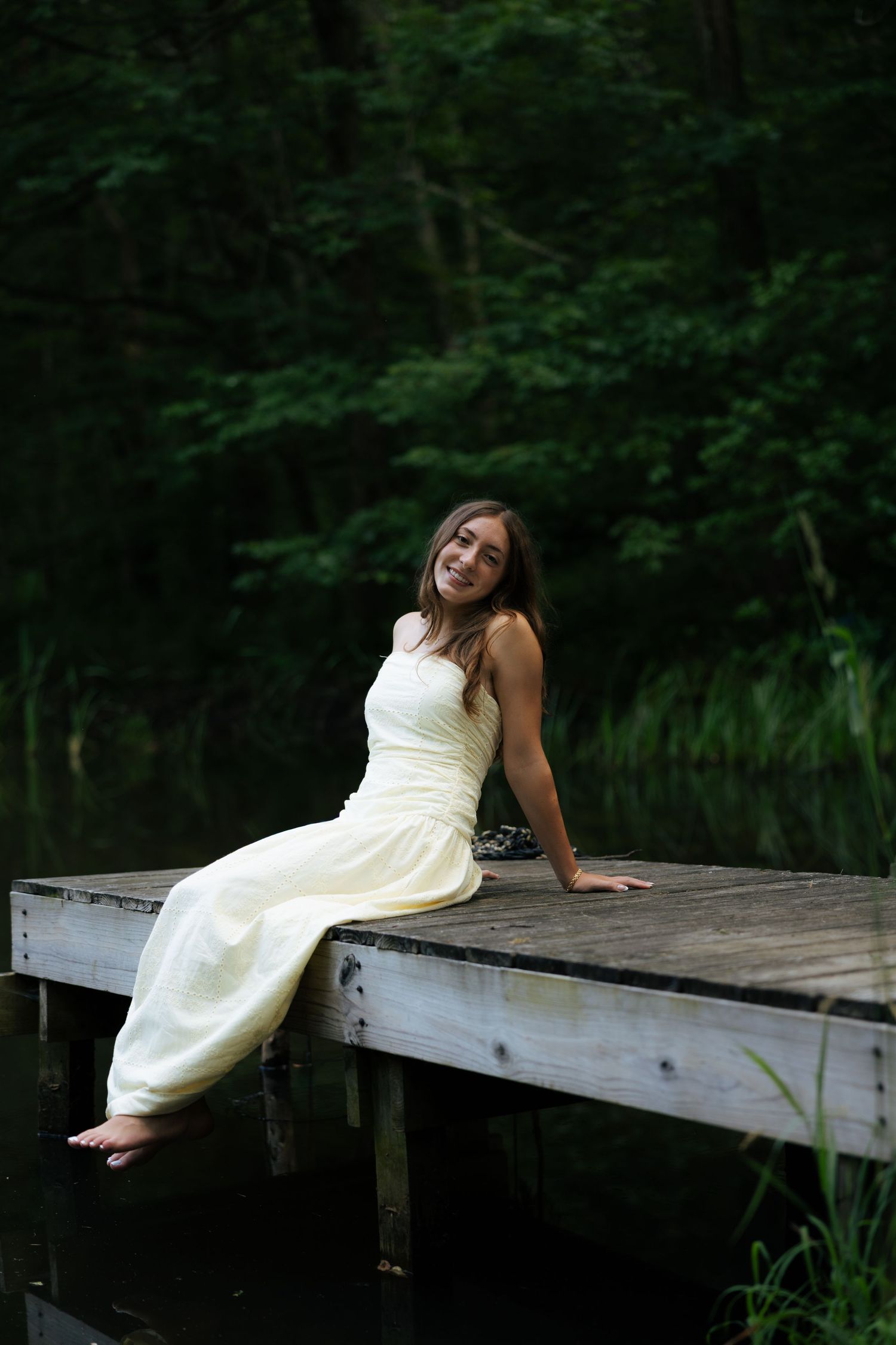 Young woman in a white dress sitting barefoot on a wooden dock by a dark forest lake, smiling.
