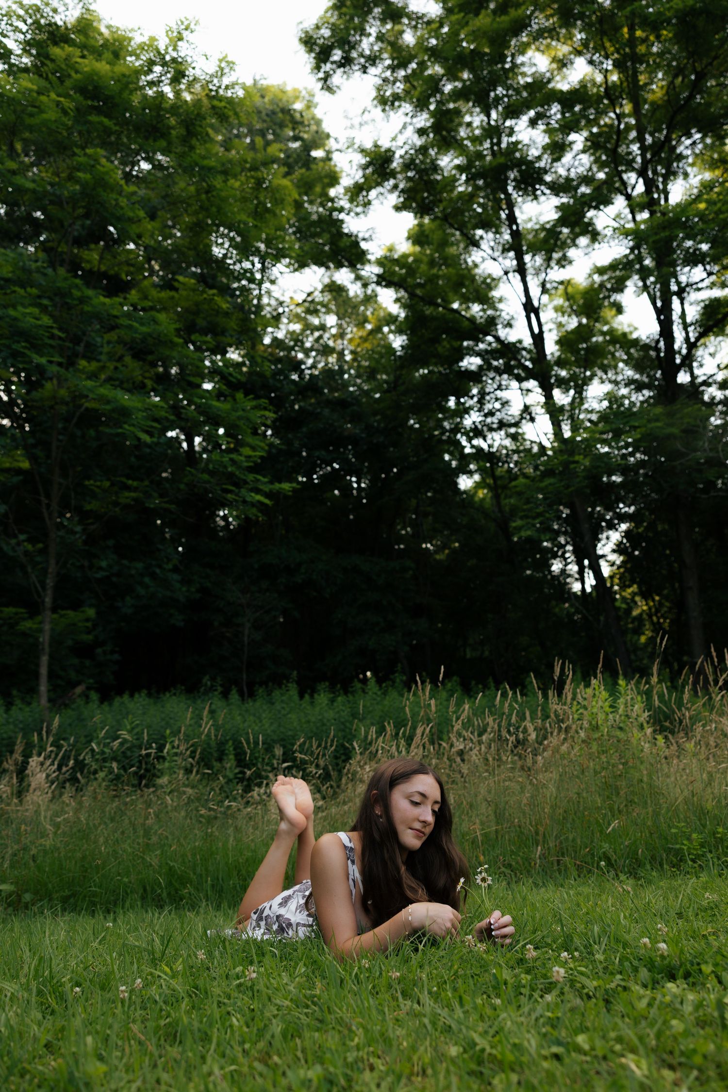 Young woman lying on grass examining white wildflowers in a lush green outdoor meadow setting.