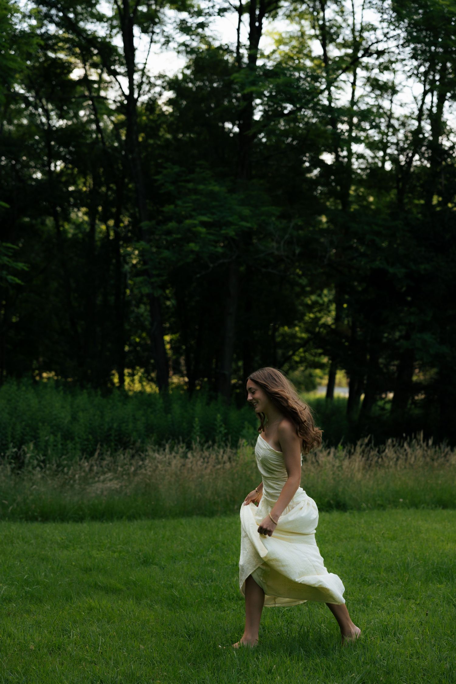 Young woman in white dress twirling barefoot on lush green grass with dark forest background.