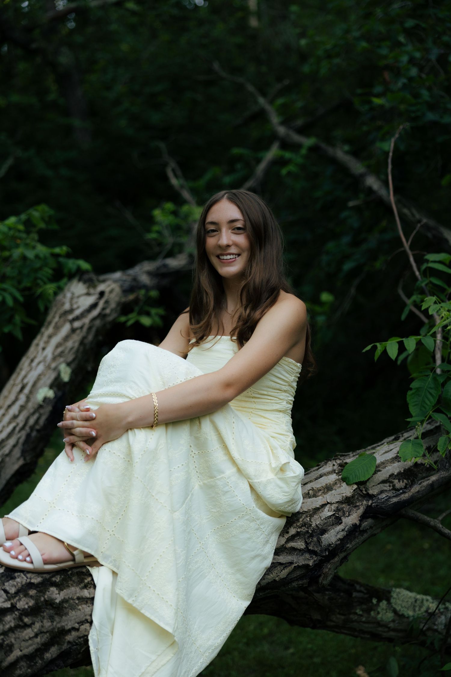 Smiling young woman in cream strapless gown sitting on fallen log in forest.