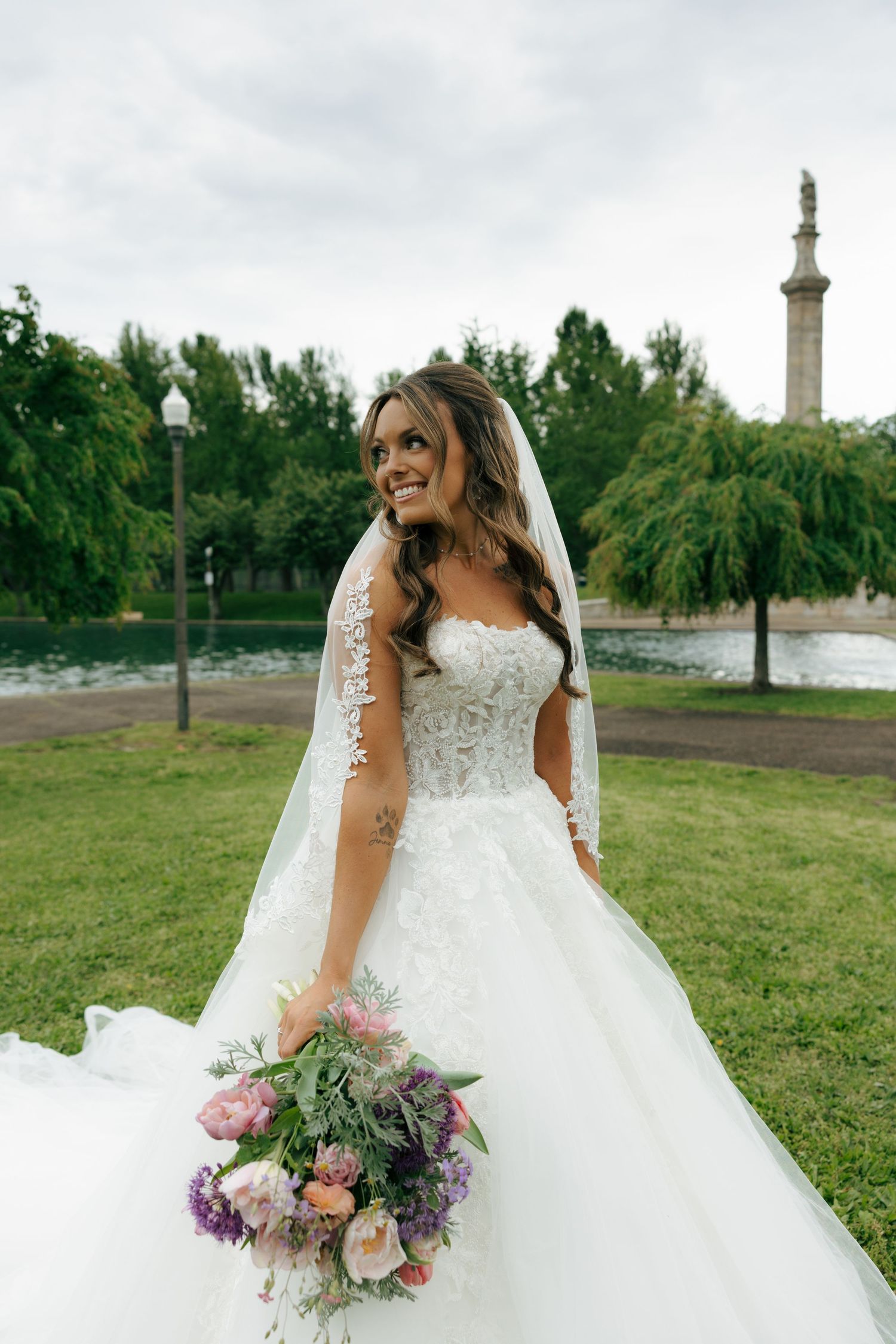 Smiling bride in lace wedding gown and veil holds colorful bouquet in a green park near a fountain.