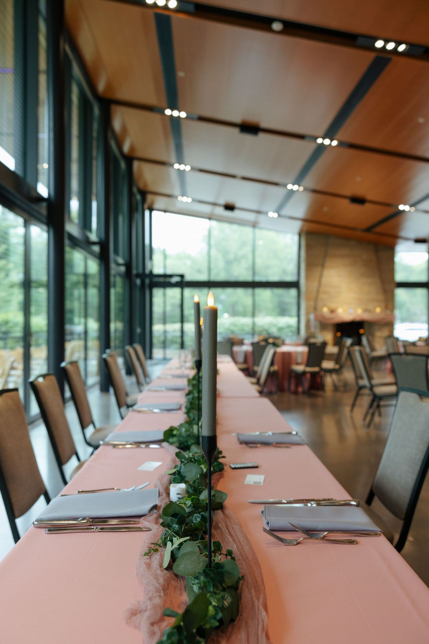 Elegant wedding reception hall with long table, pink linen, greenery garland, candles, and floor-to-ceiling windows.