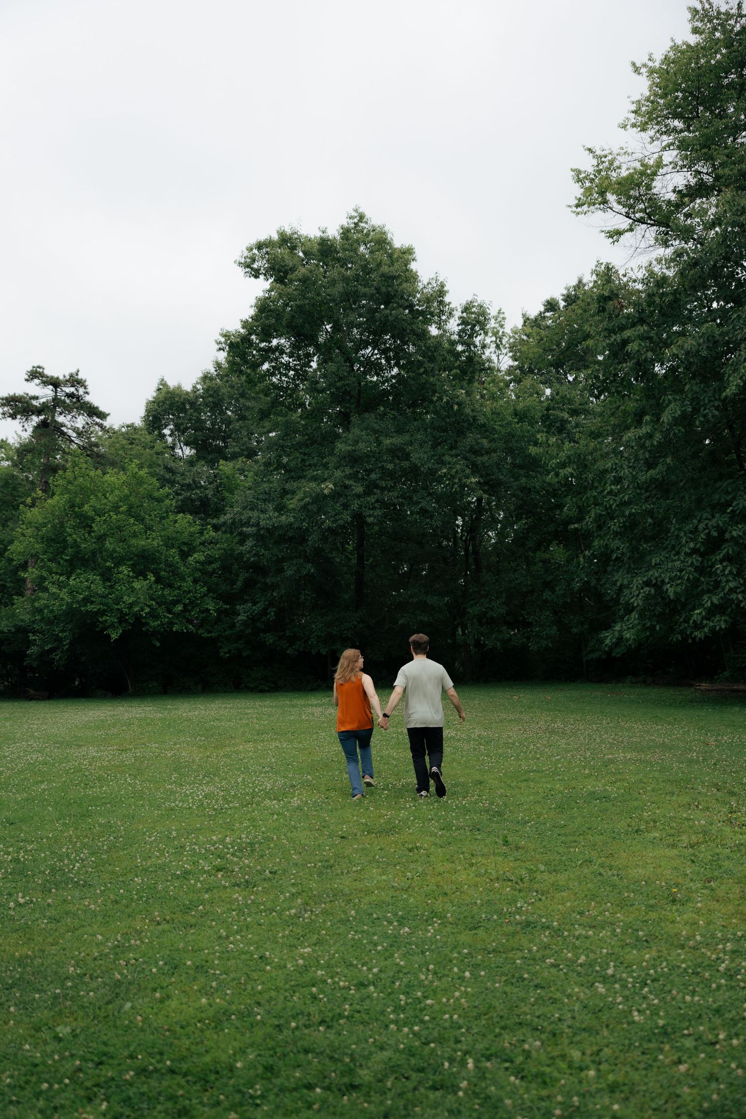 Couple walking hand in hand across a green meadow toward a dense tree line.