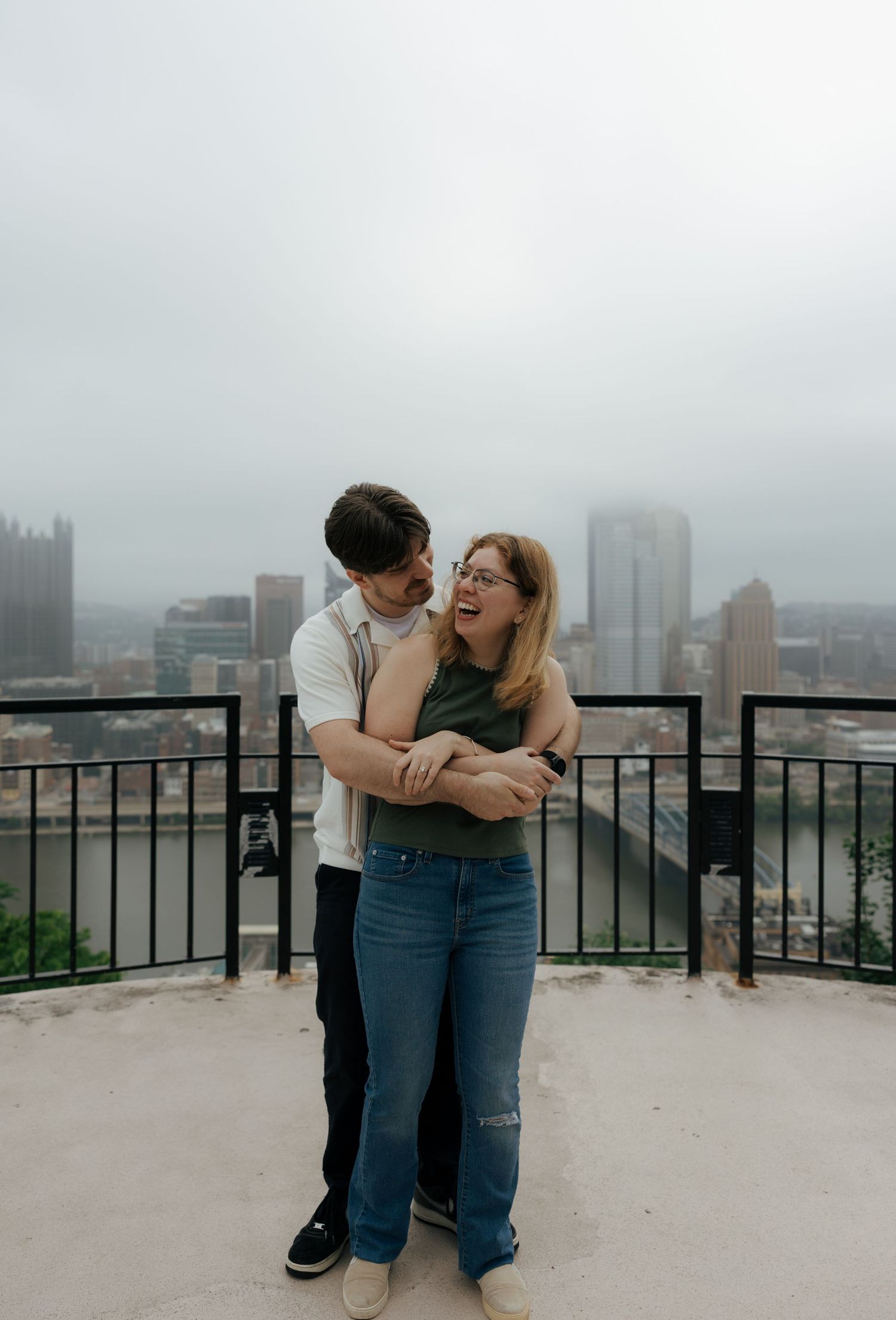 A laughing couple embraces on a scenic overlook with the Pittsburgh city skyline visible on a cloudy day.