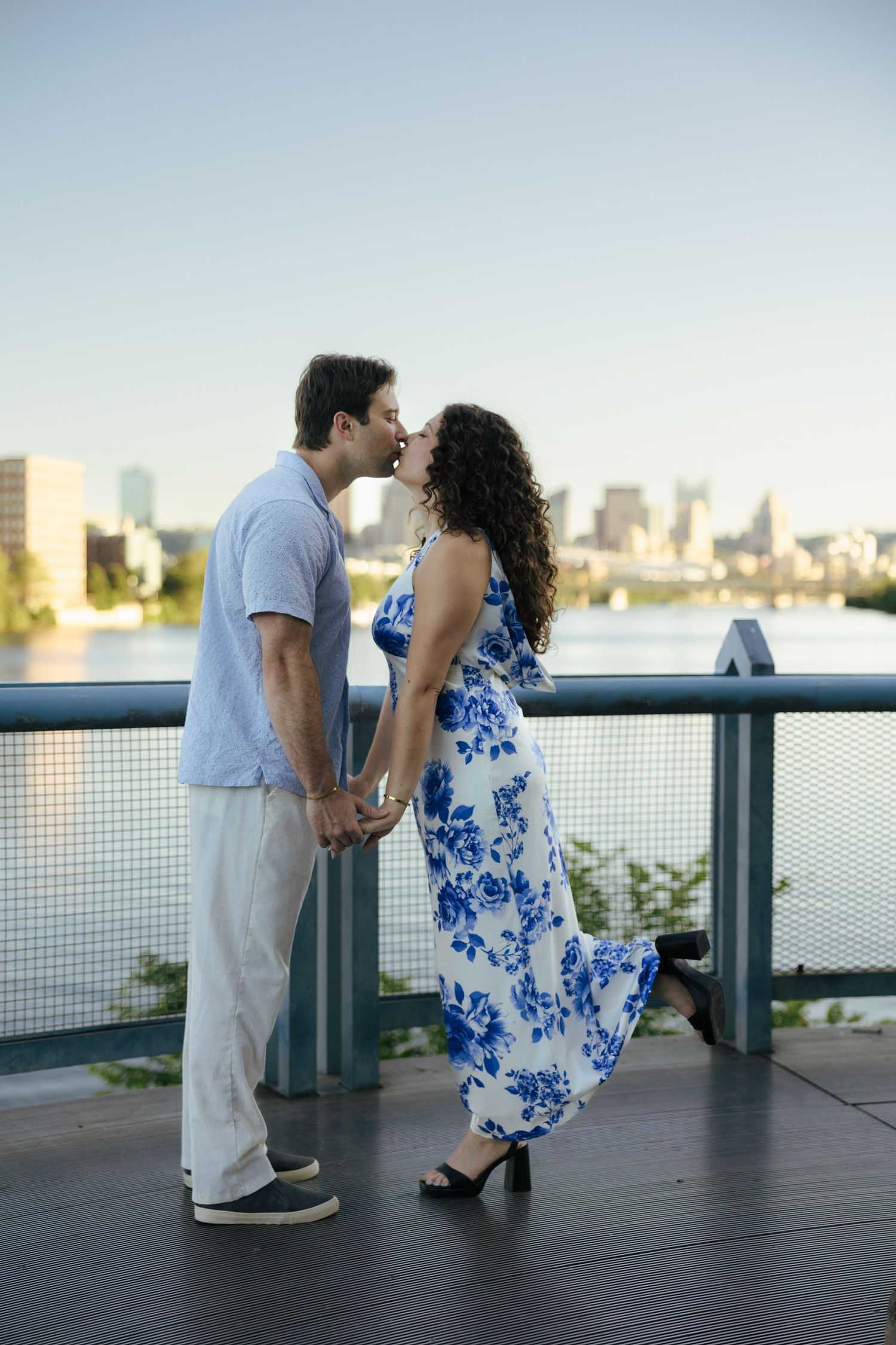 Couple sharing a romantic kiss on a waterfront bridge, woman wearing a blue floral dress.