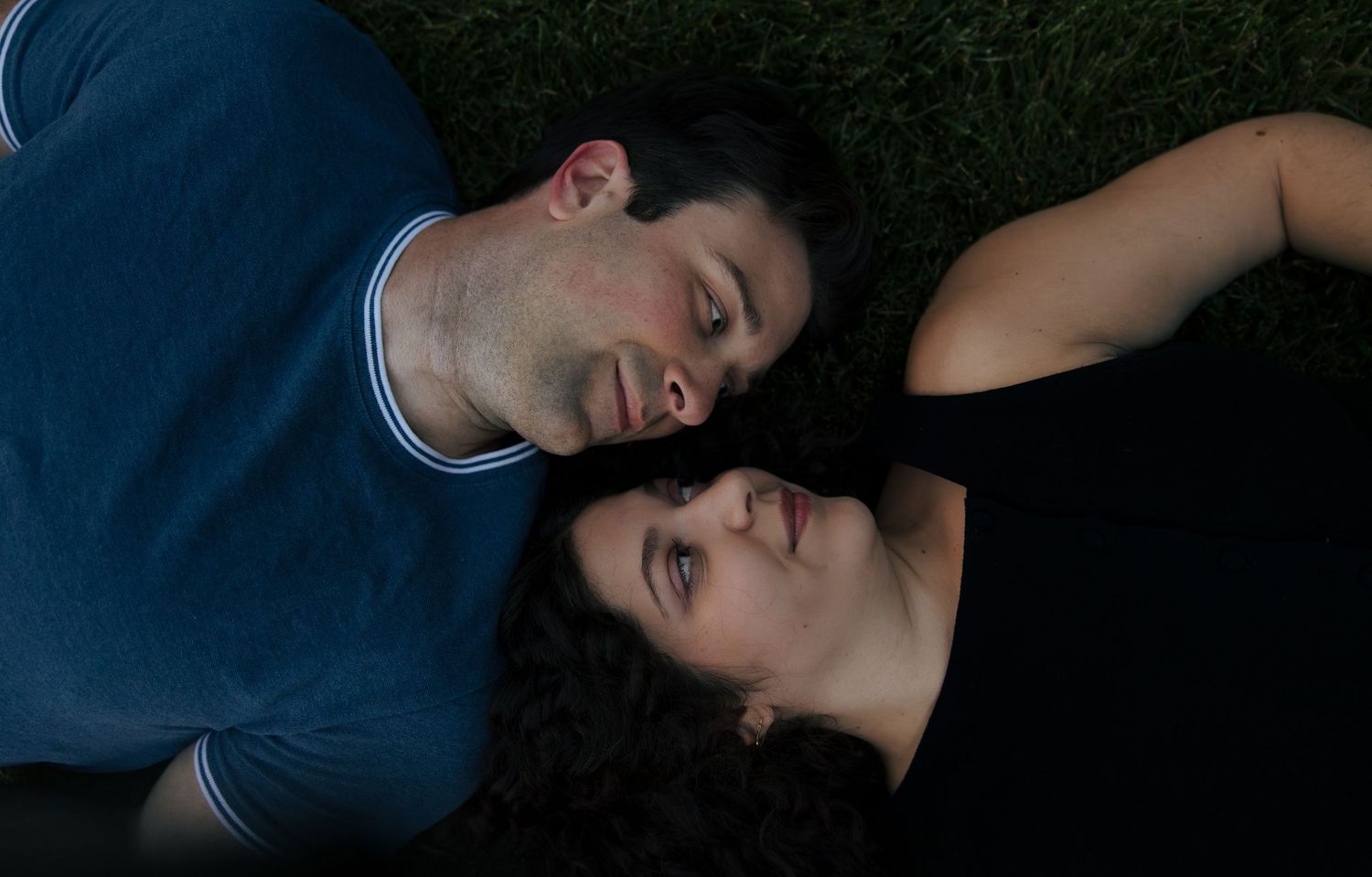 A couple lies on grass gazing at each other tenderly in a romantic overhead portrait shot at dusk.
