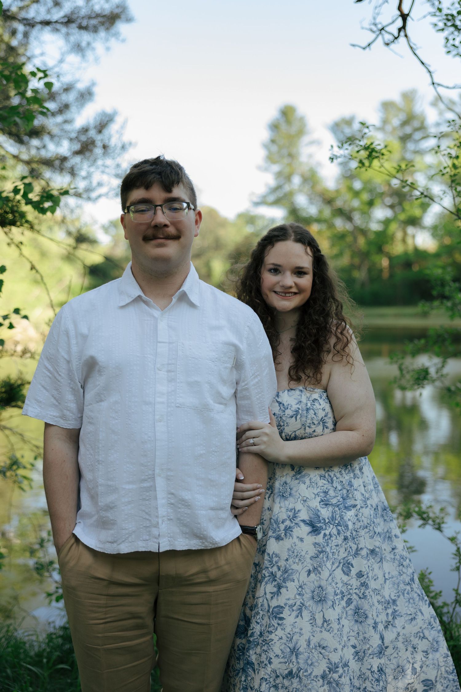 Couple posing by a serene pond surrounded by lush green trees, woman in floral dress embracing man in white shirt.