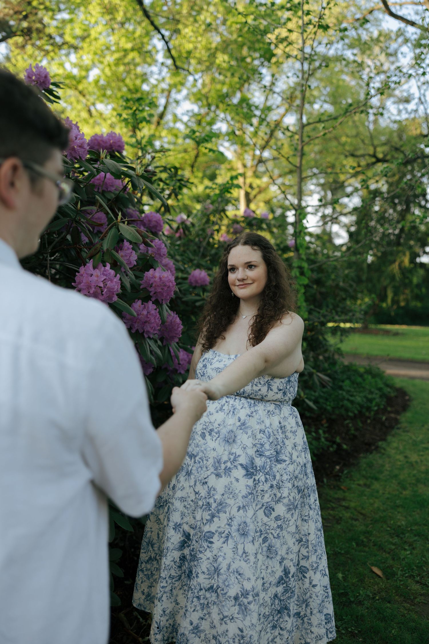 Woman in floral dress smiling at partner while holding hands near purple rhododendrons.