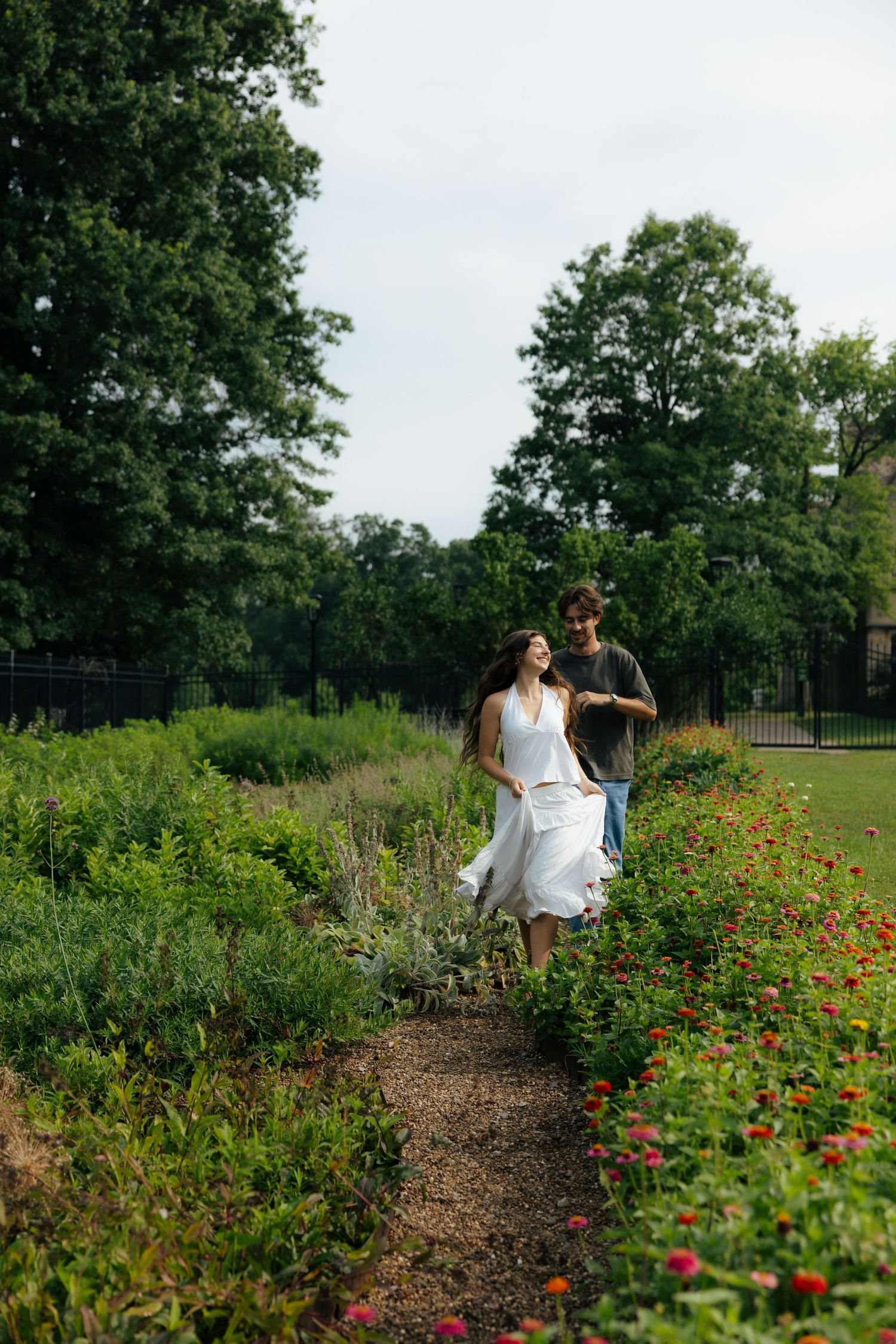 A couple strolls together along a vibrant garden path with red flowers and green trees.