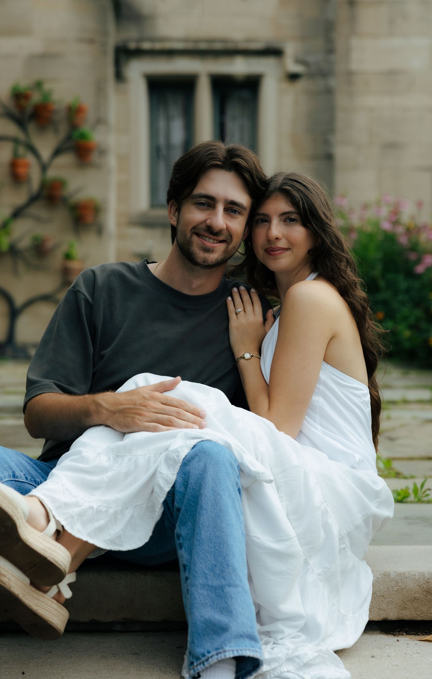 A couple sits closely on stone steps outside a historic building, the woman in a white dress leaning on the smiling man.