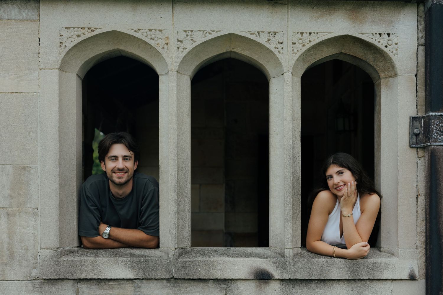 Smiling couple leans out of arched stone windows of a historic building, posing playfully for the camera.