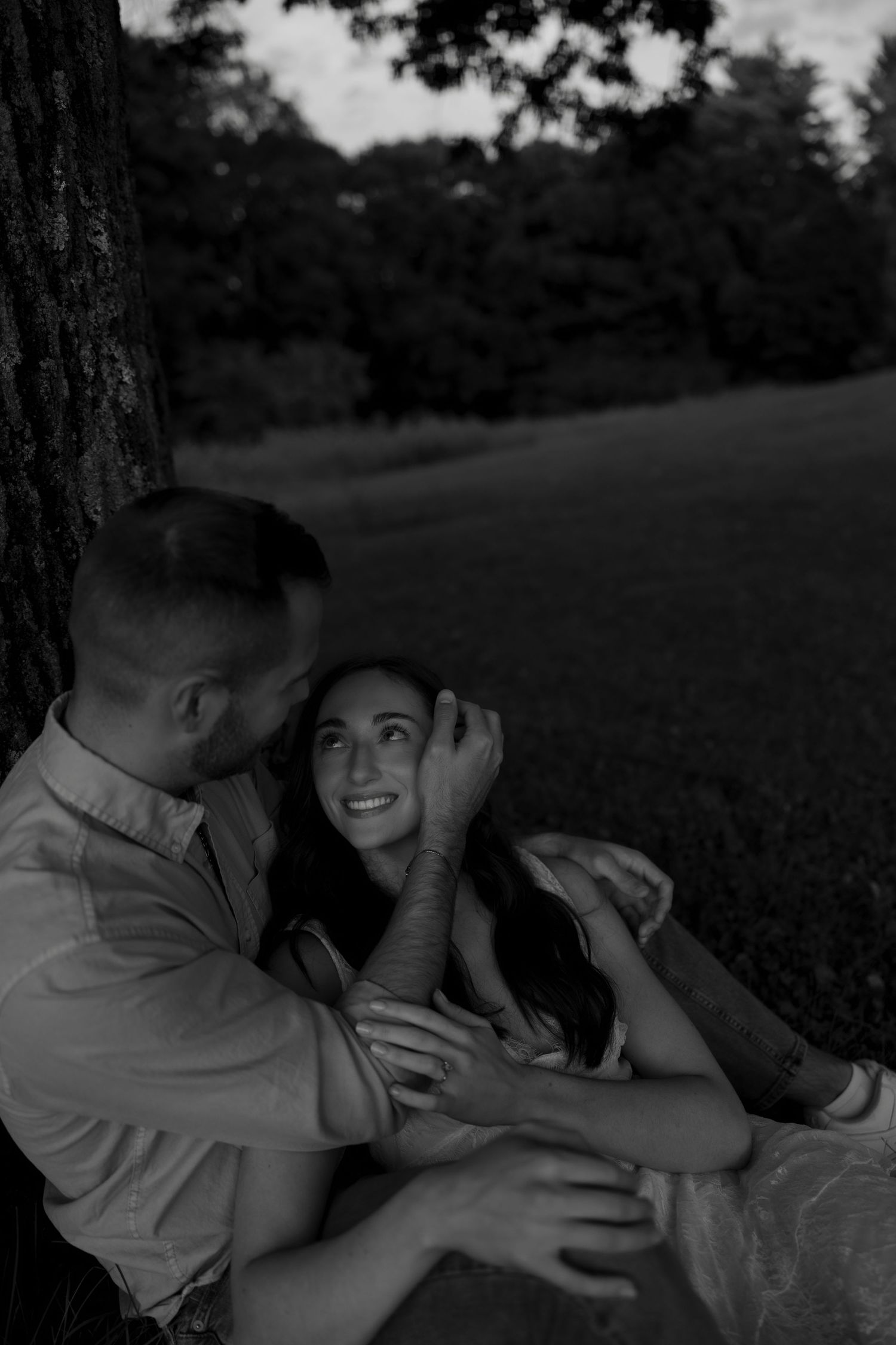 Black and white photo of a couple sitting against a tree, sharing a tender romantic moment outdoors.