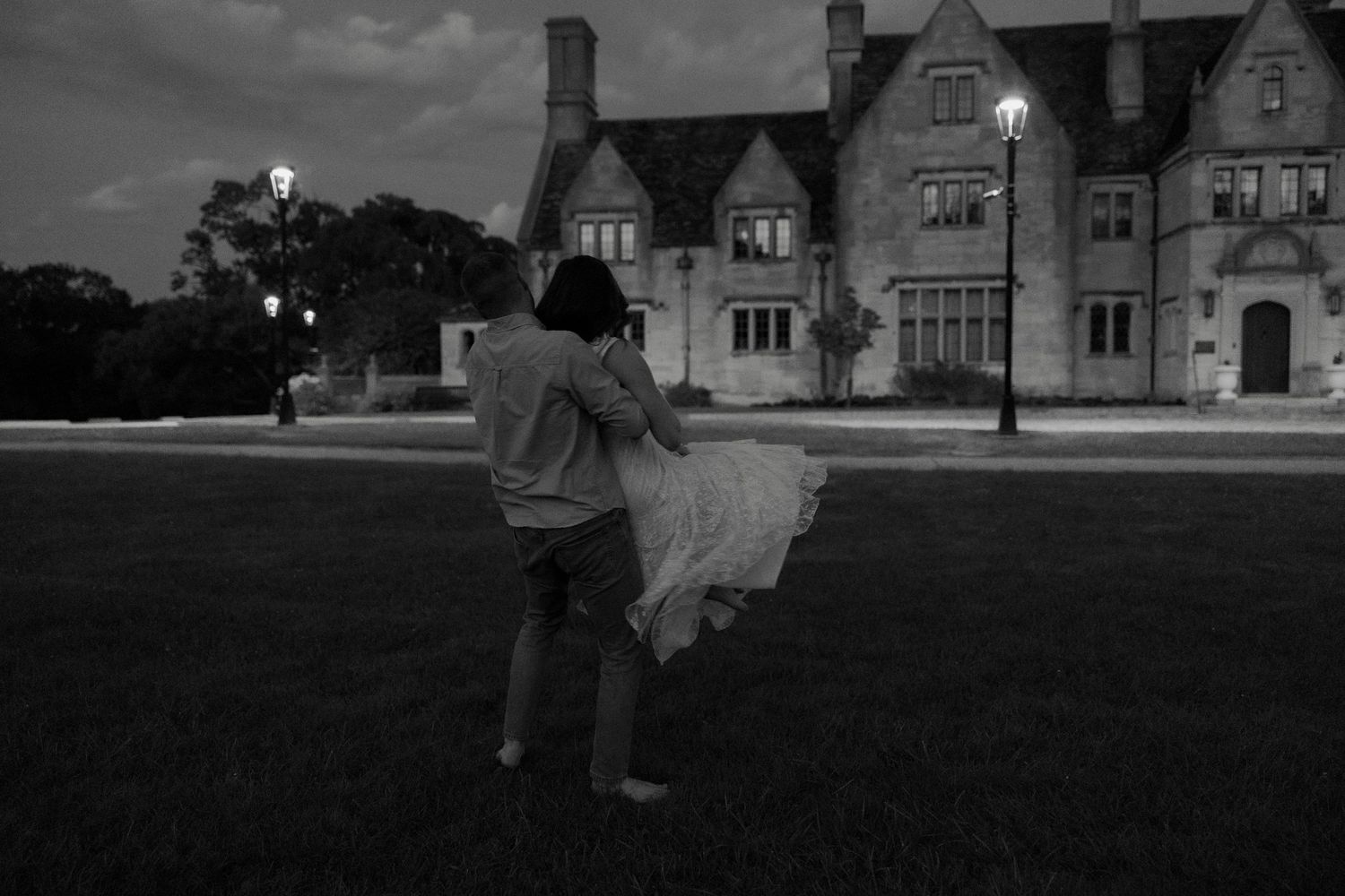 Black and white photo of couple dancing on lawn before an elegant estate at twilight.