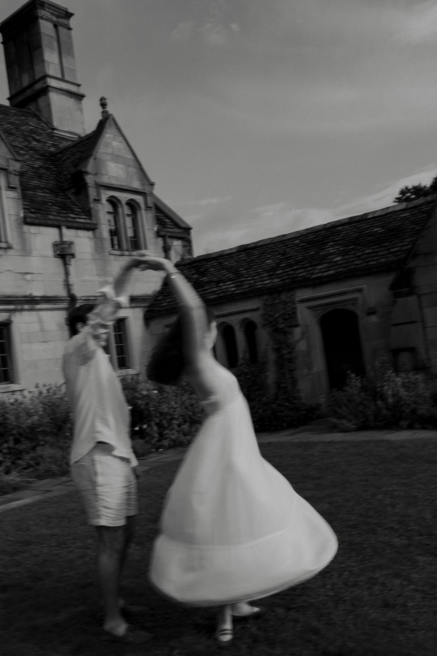 Black and white photo of a couple dancing and twirling outside a historic stone building.