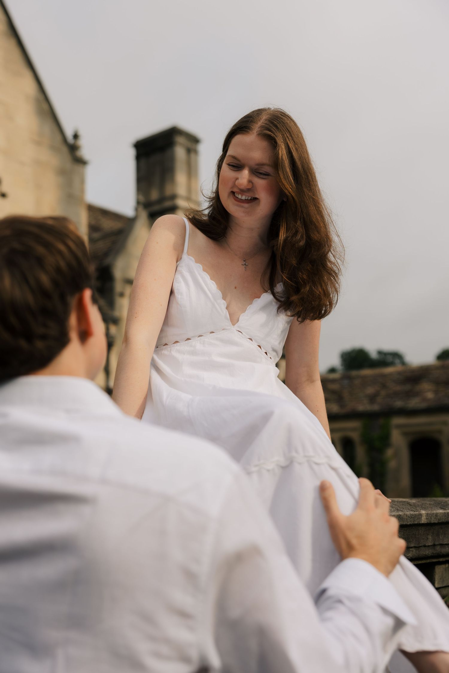 Smiling woman in white sundress sitting on stone balustrade holding hands with her partner.